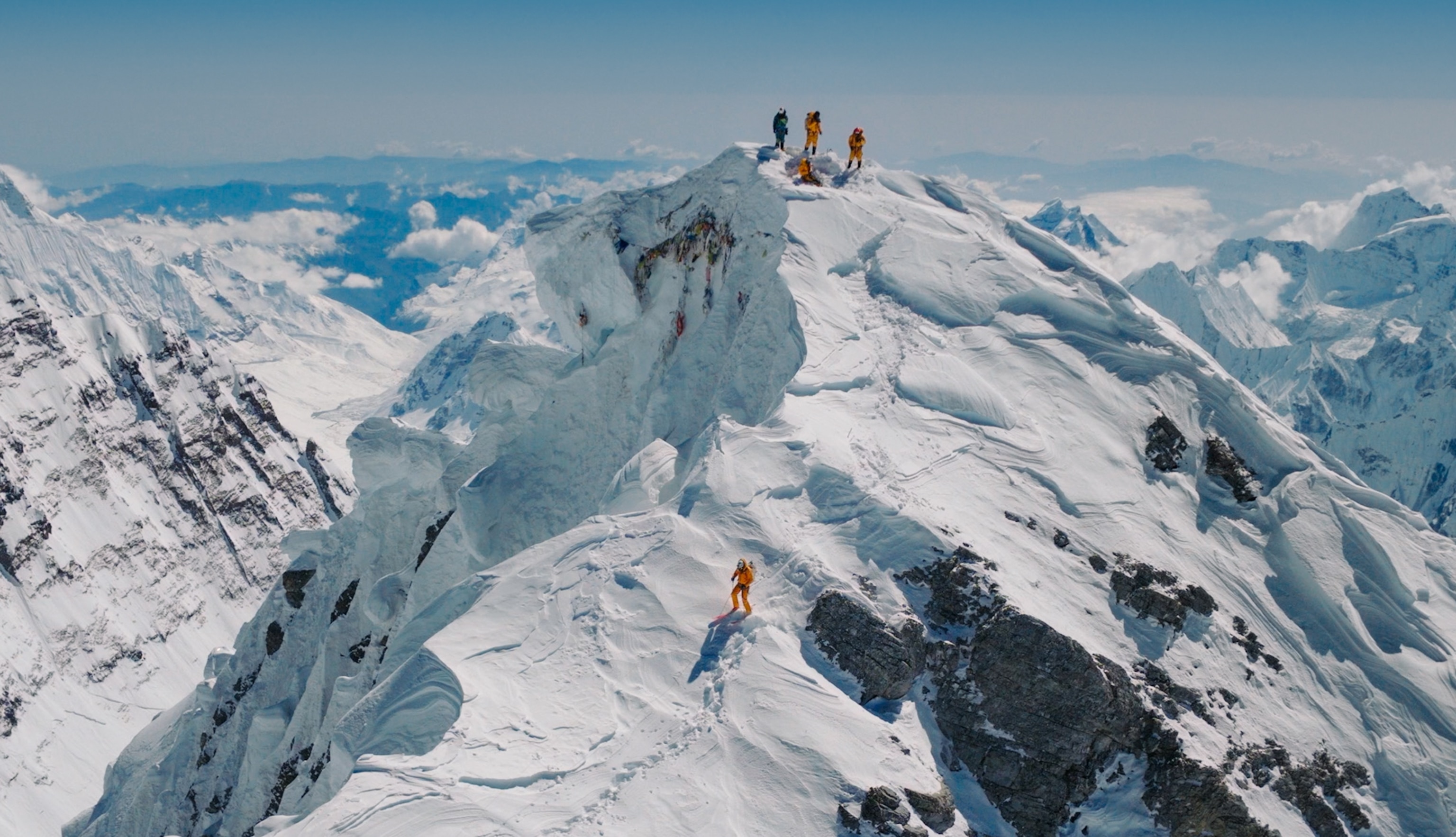 Snowy mountain landscape with 4 skiers atop the mountain. 3 wear yellow snow outfits and another wears a dark one. Another separate skier trails down the side of the snowy mountain