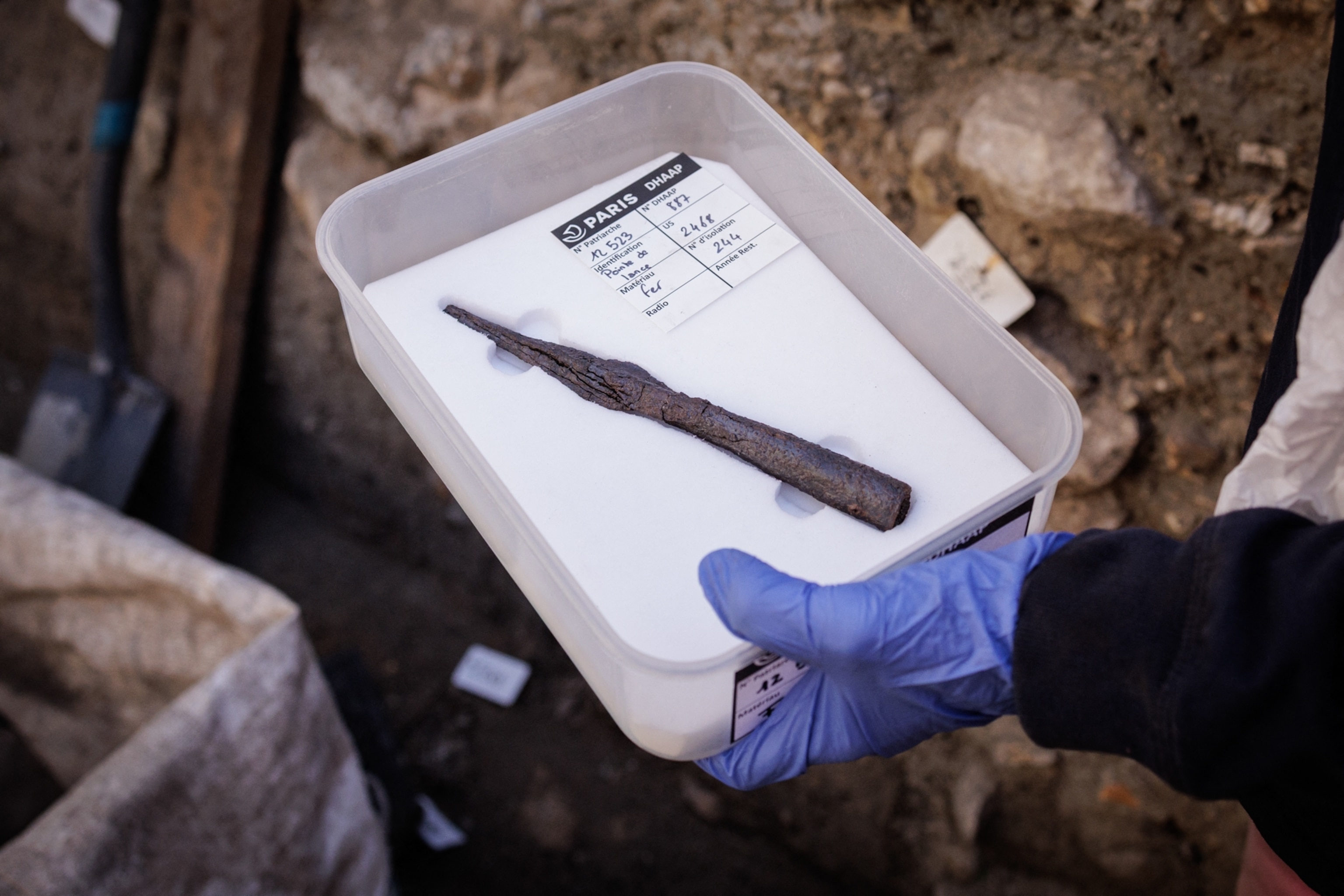 A gloved hand holds a labeled container displaying a corroded iron spearhead or metal tool recovered during an archaeological excavation.