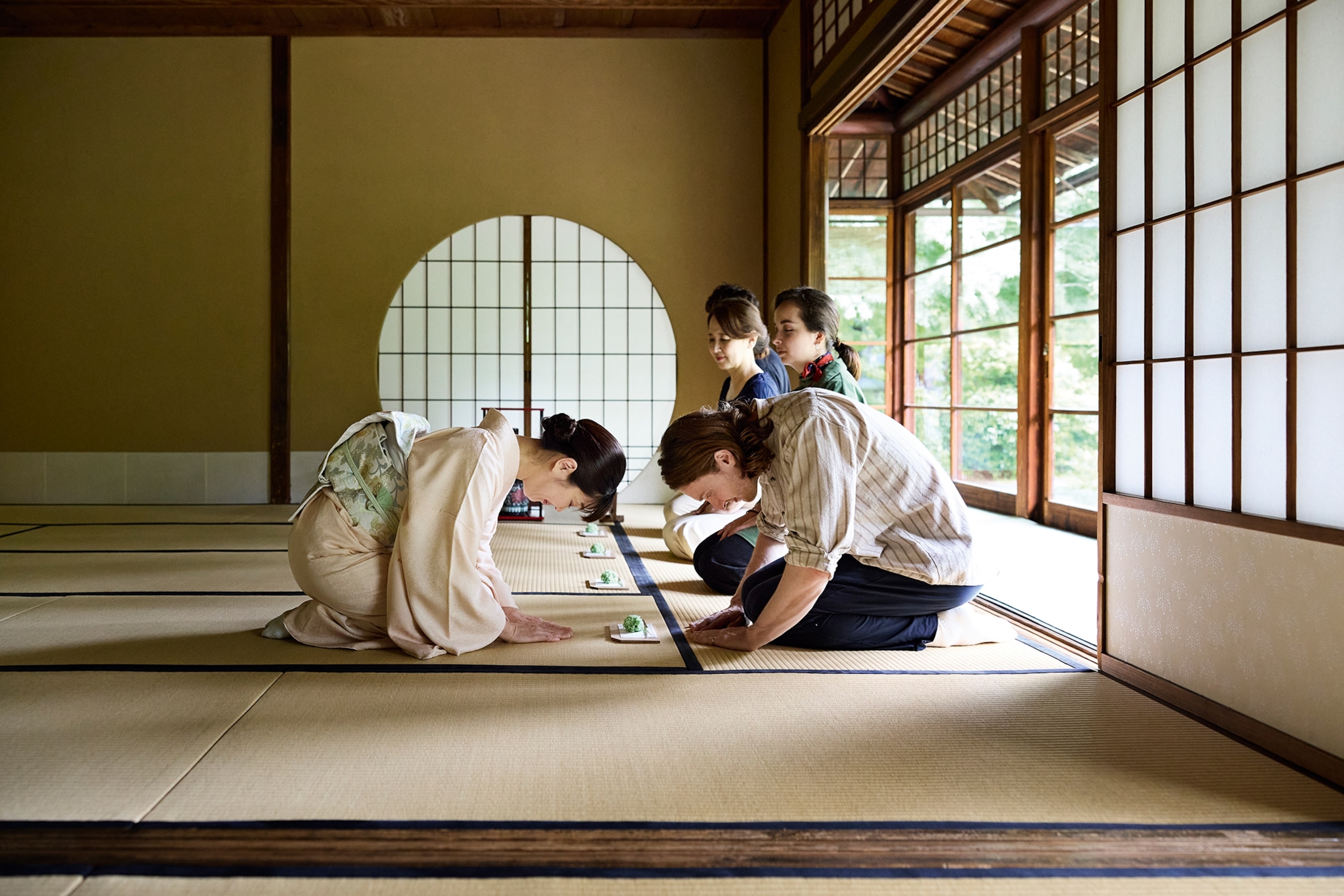 A seated woman in kimono bowing to an equally bowing foreigner in a tea room with wooden windows and three more visitors sitting in line.