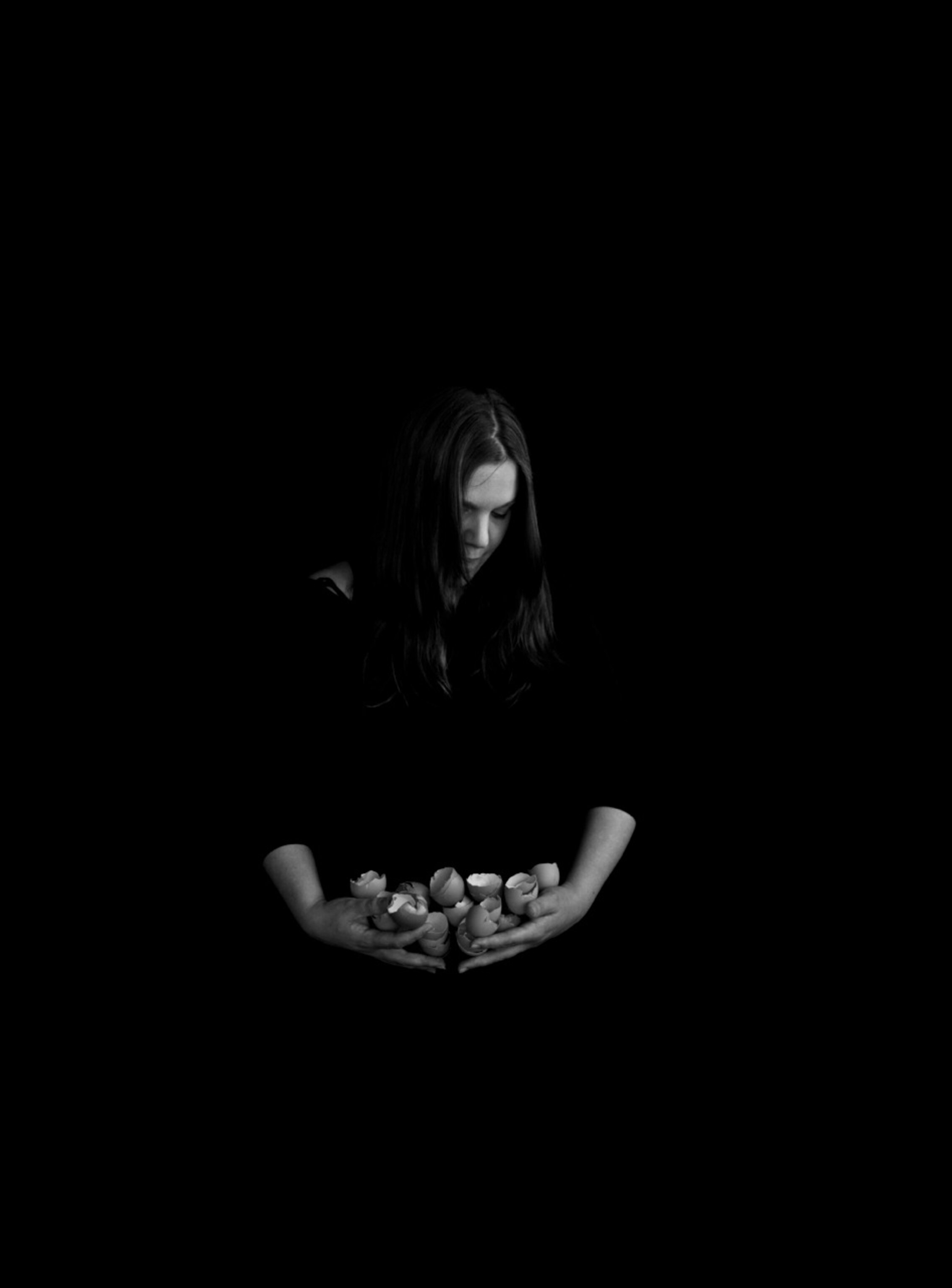 Black and white photo of a woman gazing down at the shells of eight broken eggs that she cradles in her arms