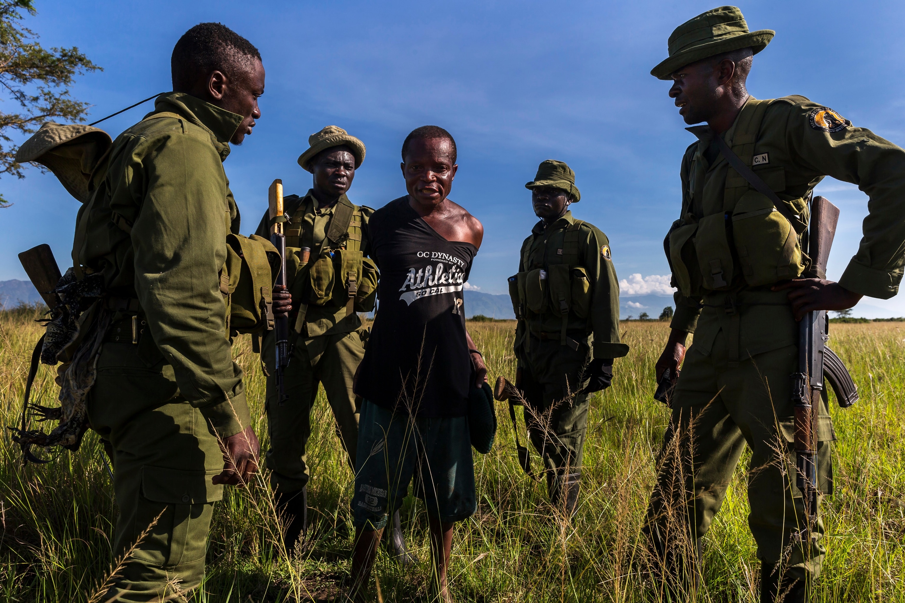 a man stands with his arms behind is back as rangers surround him in the grass