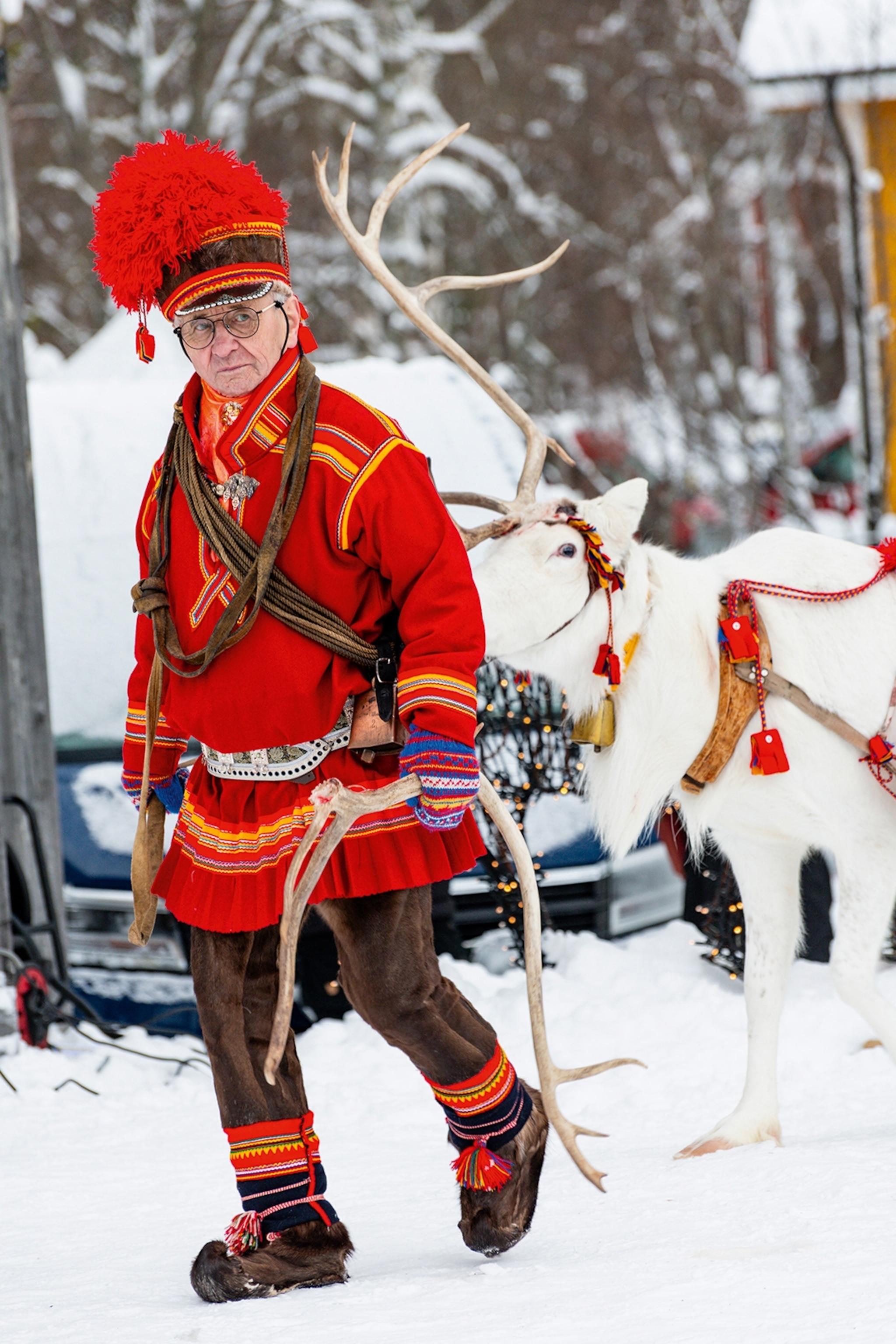 An elderly man dressed in traditional, embroidered winter clothing and a tassel-topped hat, guiding a reindeer through a winter landscape.