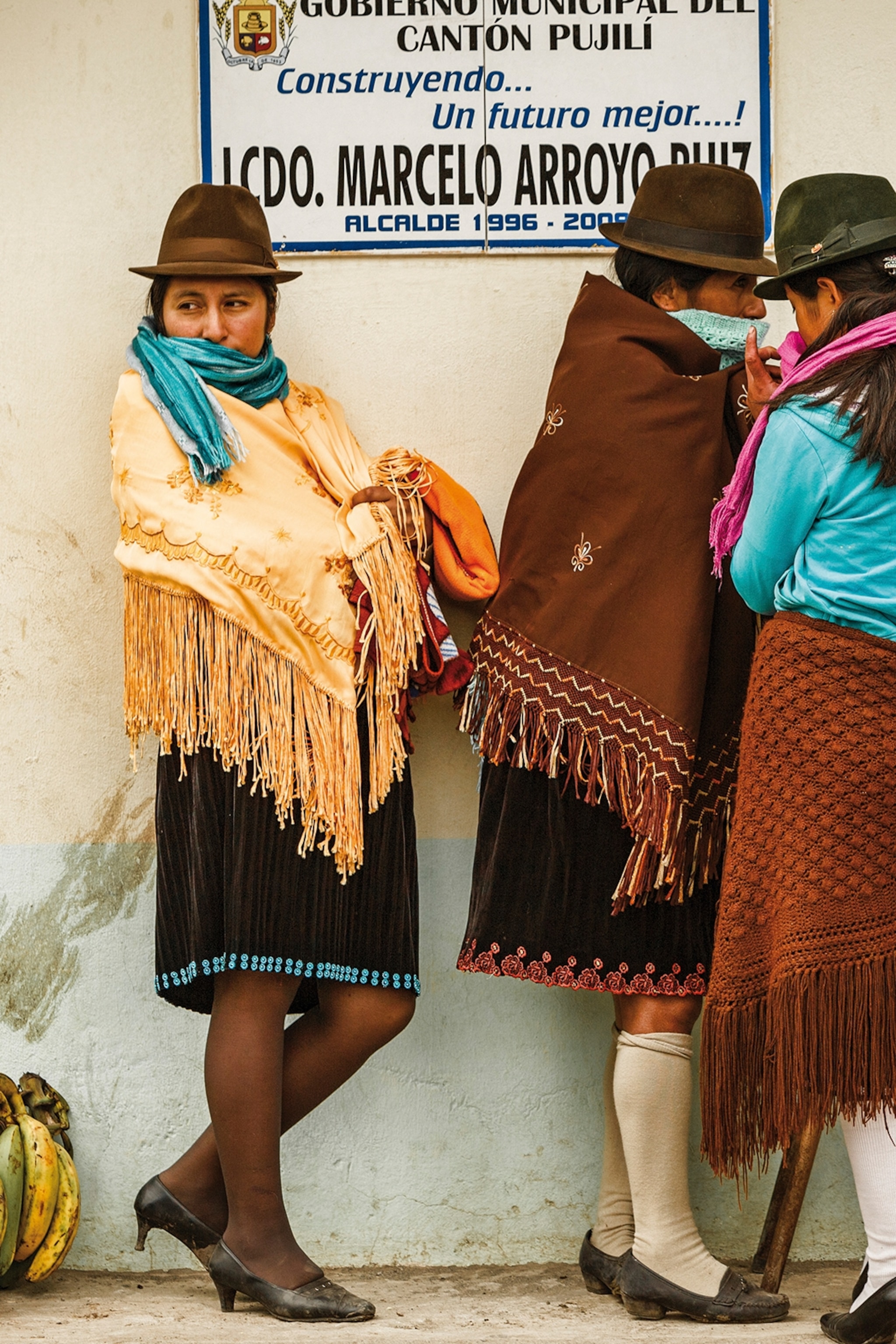 Three local women wearing bowler hats and shawls, leaning against a wall on a market.
