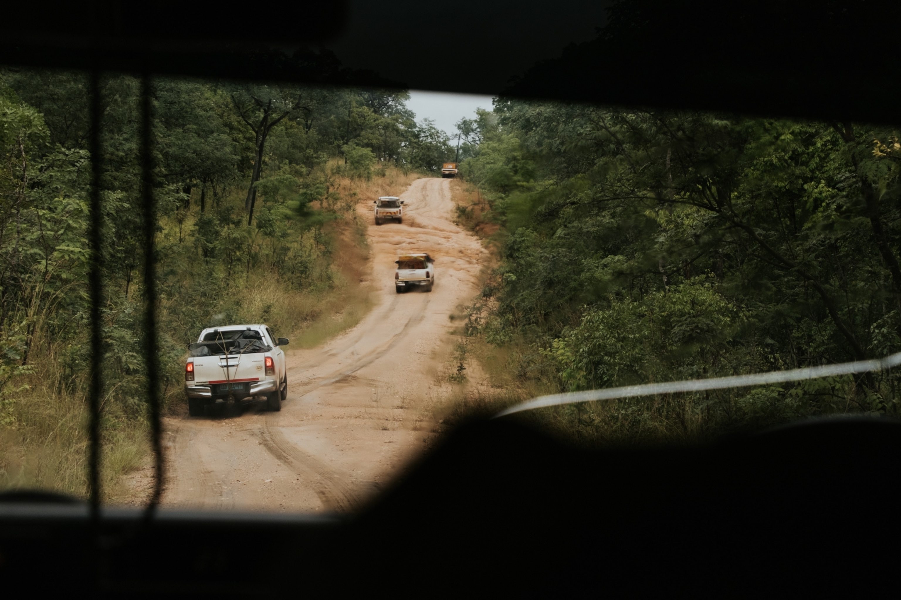 Car convoy on a rugged dirt road winds through dense green forest, viewed from inside another vehicle
