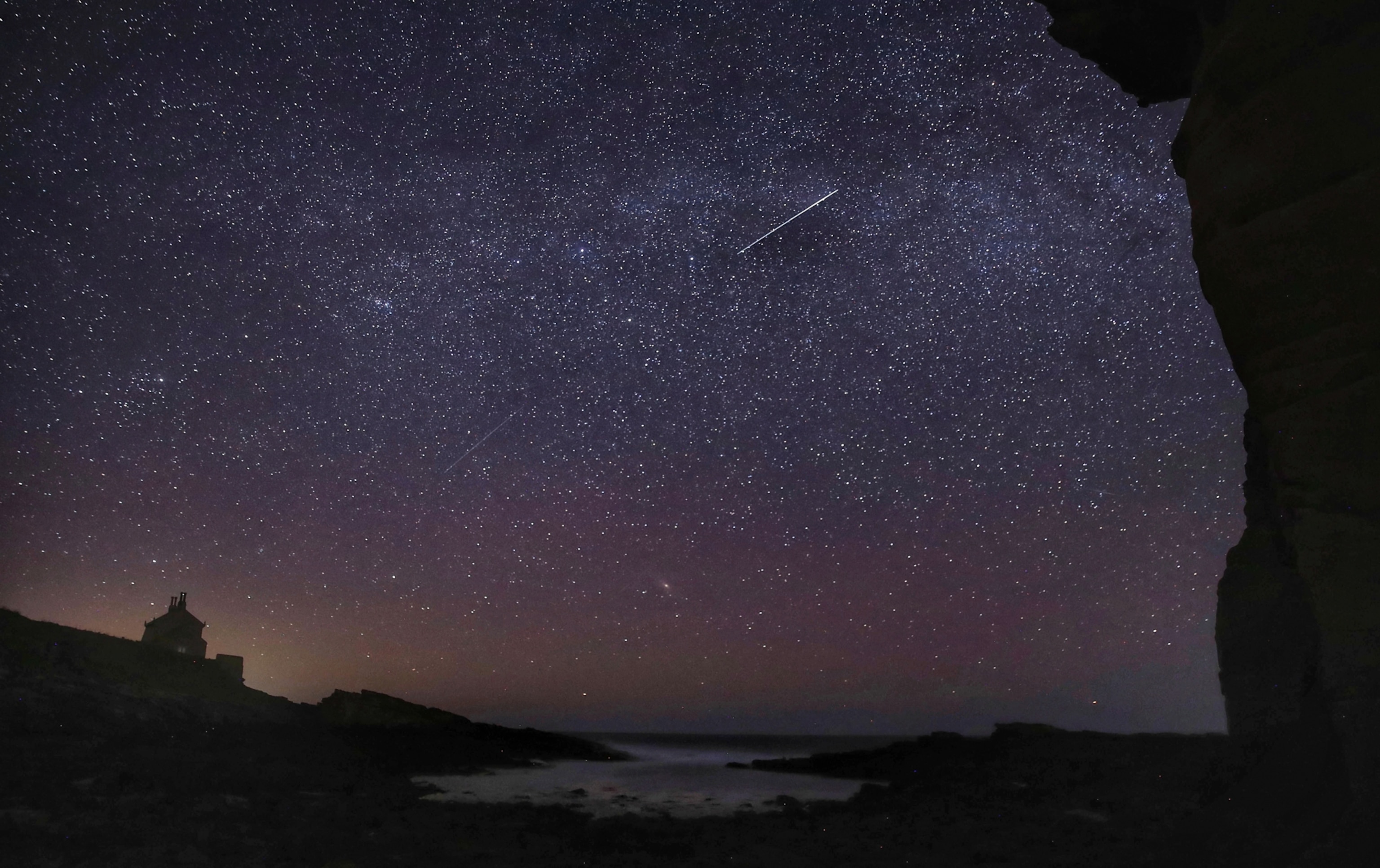 Starry night sky with a visible shooting star over a rocky coastline. Silhouette of a house on the left