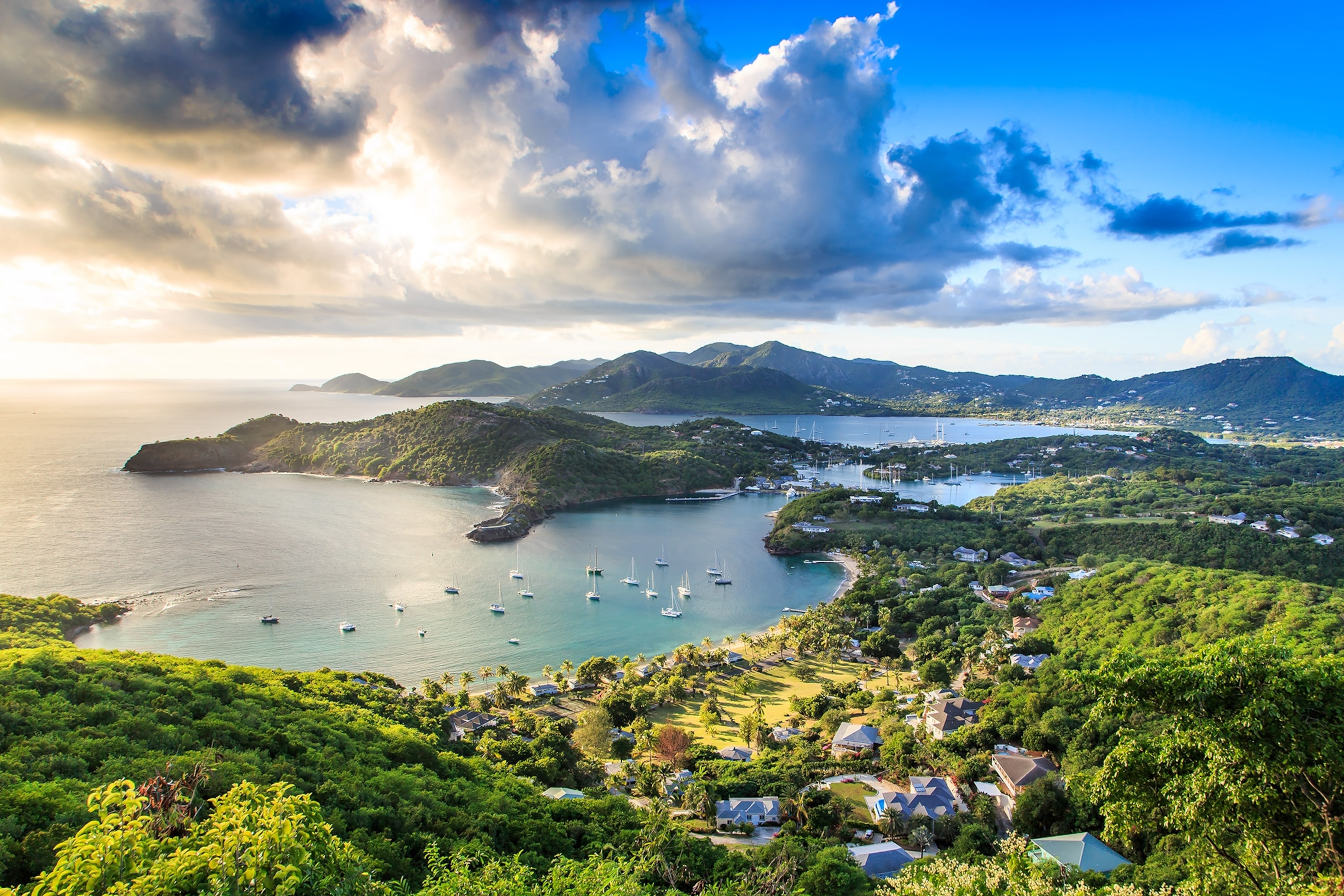 Aerial of a bay surrounded by land with lush greenery and dotted with small buildings. Sun pierces through dramatic clouds above in a blue sky