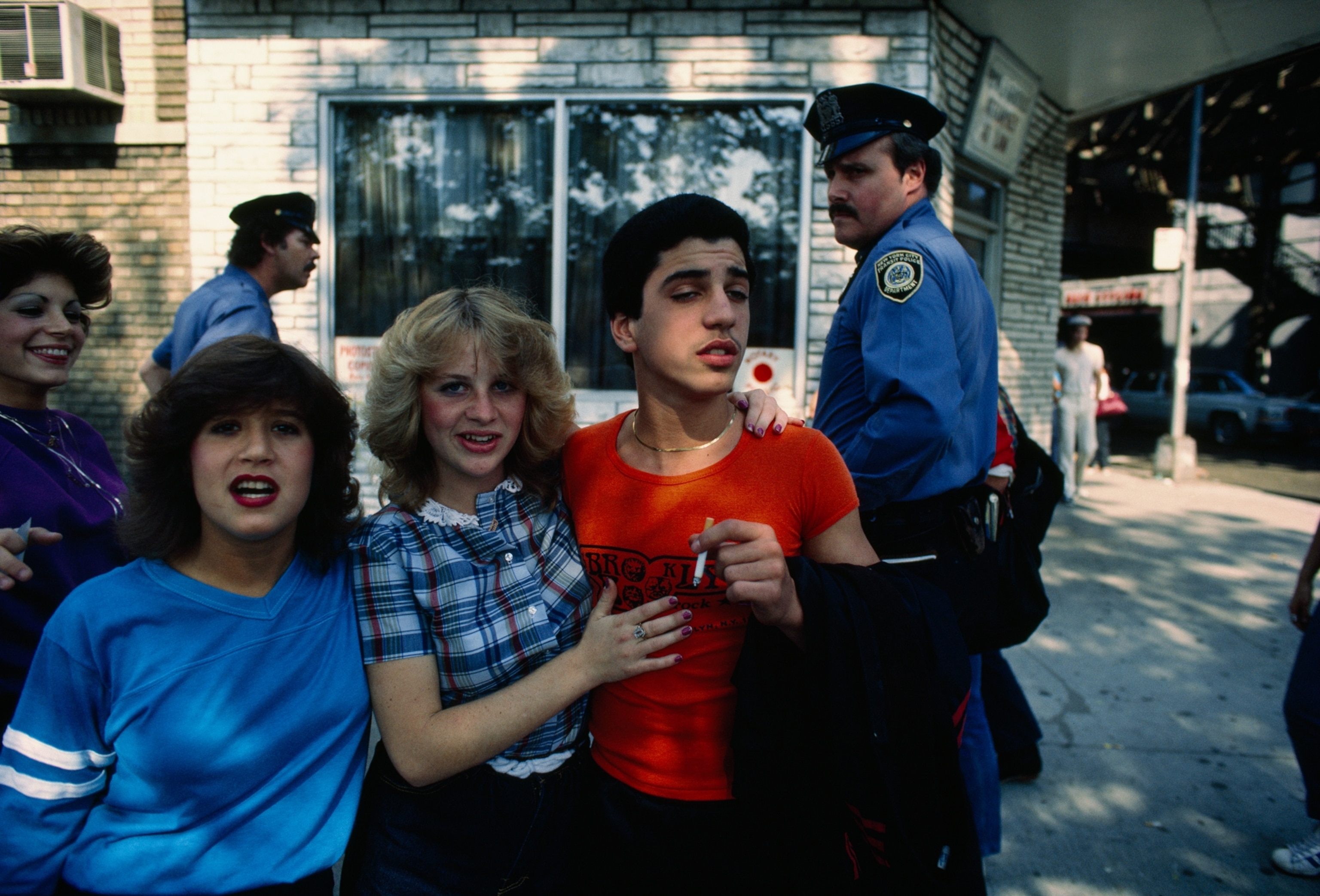 Eighties Photos - Three teenagers mug for the camera as two policemen look on, Bay Ridge, Brooklyn, 1983.