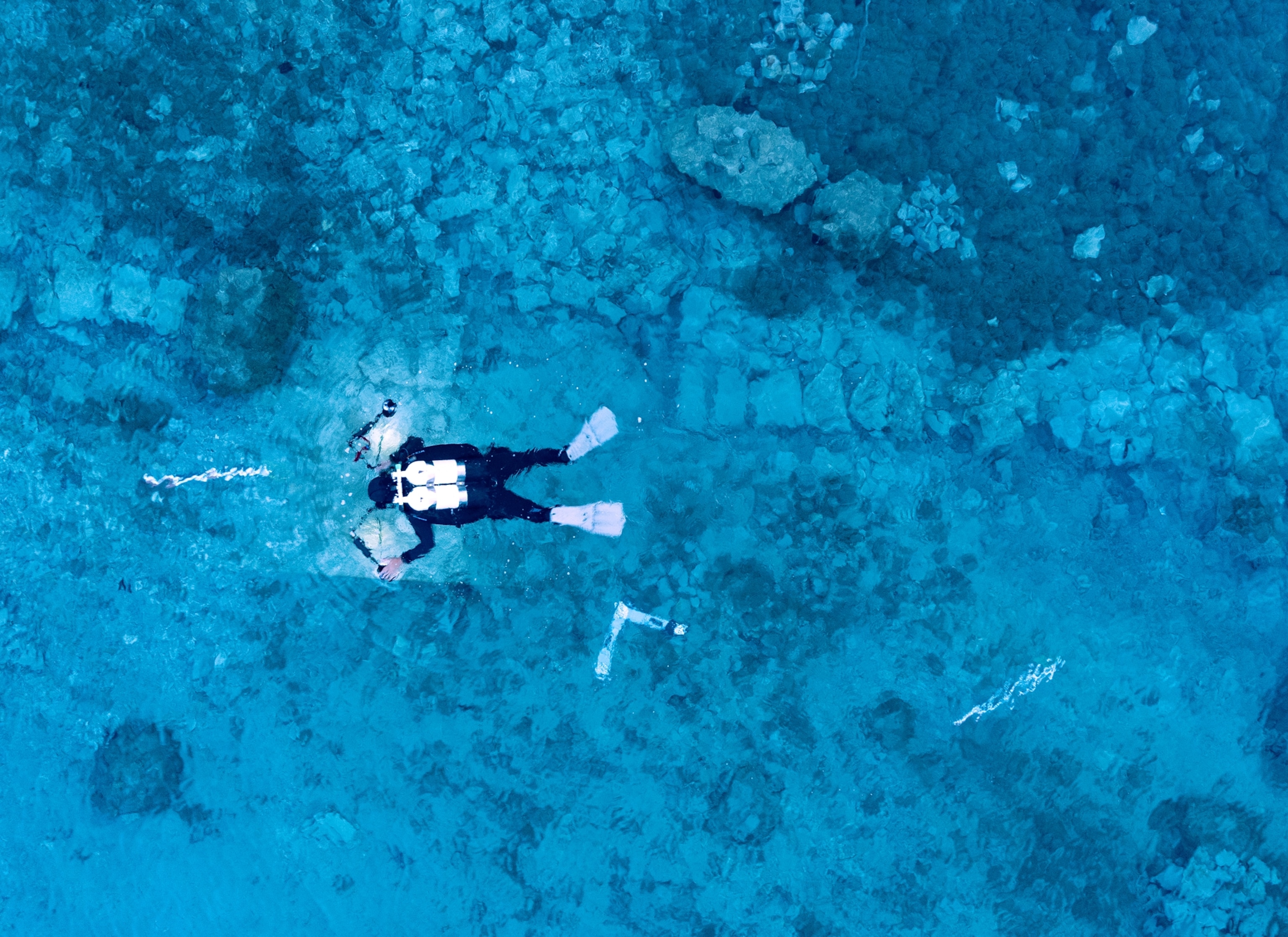 The full body of a diver using an underwater camera and wearing a black wet suit, grey flippers, and a white oxygen tank can be seen against the background of very blue water.