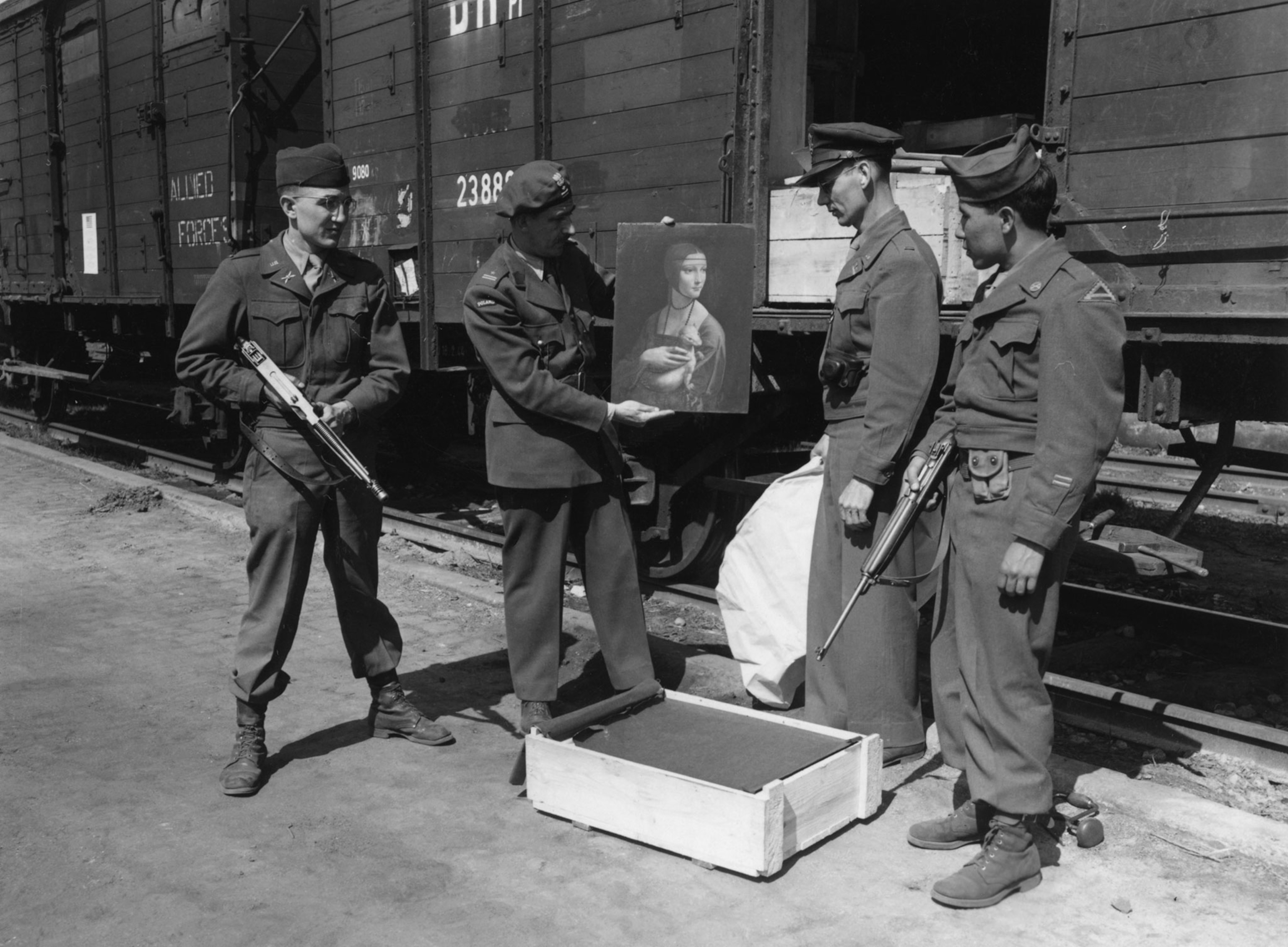 Three men in uniform stand together in a black and white photograph. They are holding a painting in front of a train not currently in motion.
