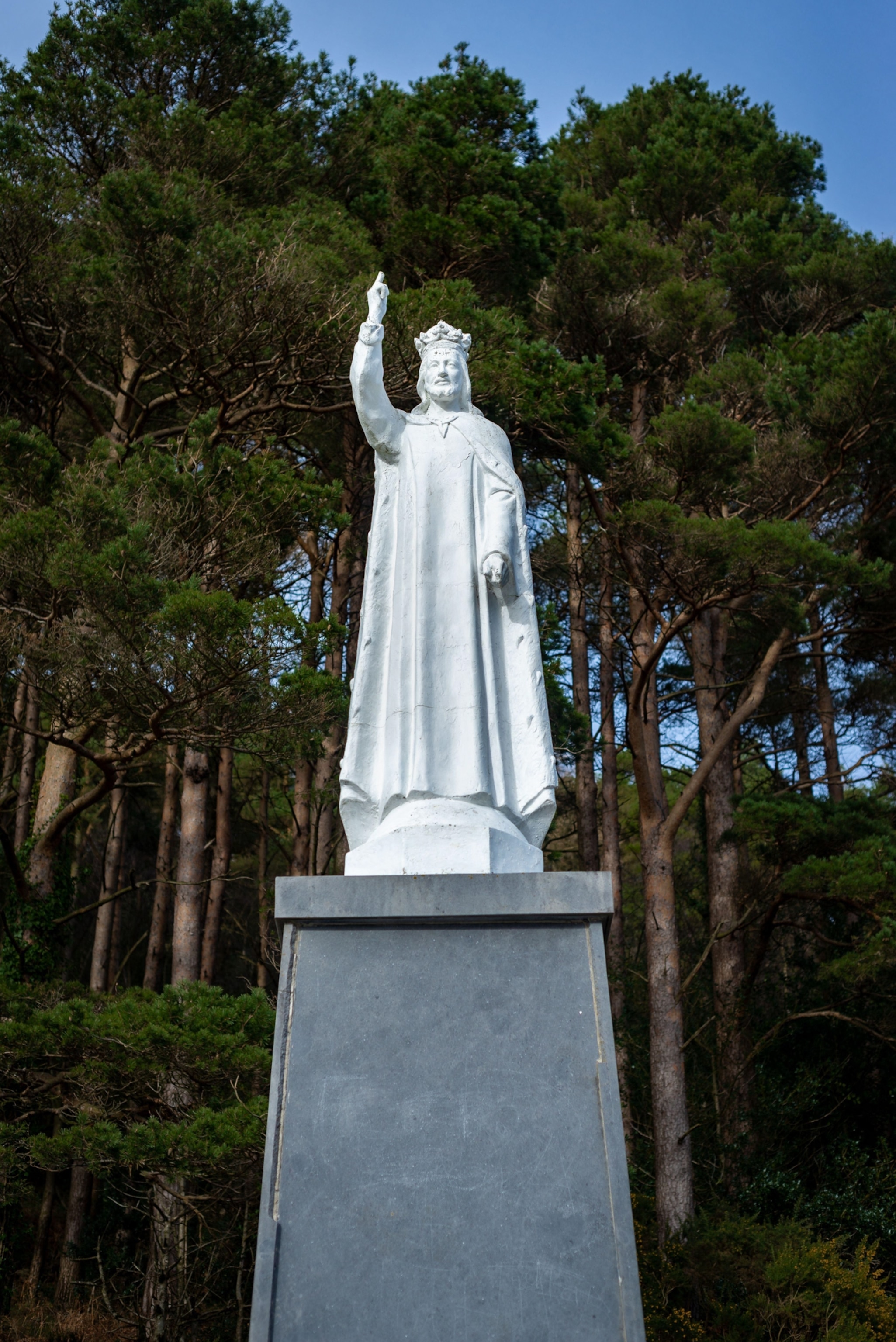 A white statue of Jesus standing atop a grey granite column with pine trees in the background