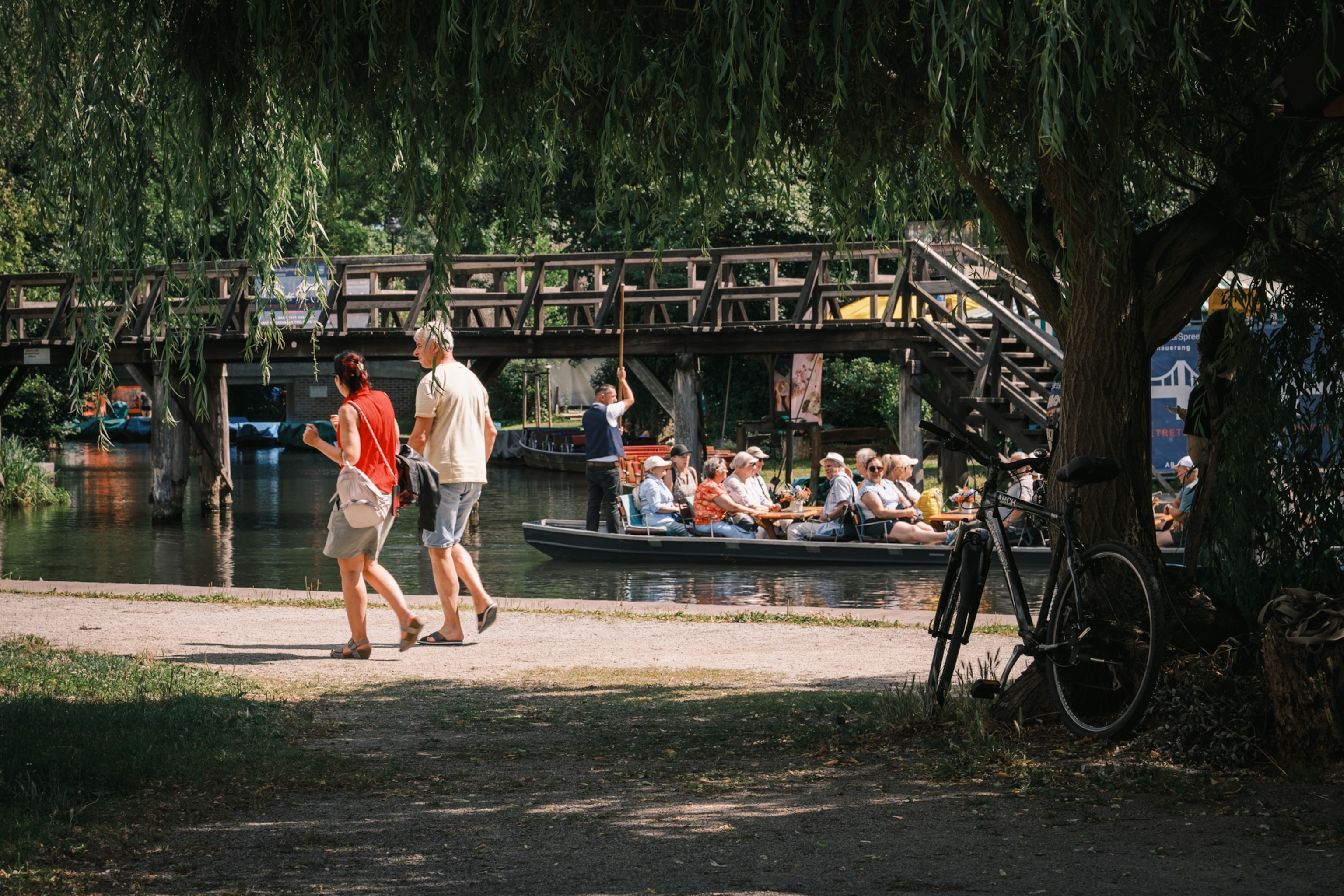 A scene in Lübbenau at the “Großer Spreewaldhafen” on the Hauptspree. Visible in the image are pedestrians, a parked bicycle, a traditional Spreewald boat, and a pedestrian bridge in the background.