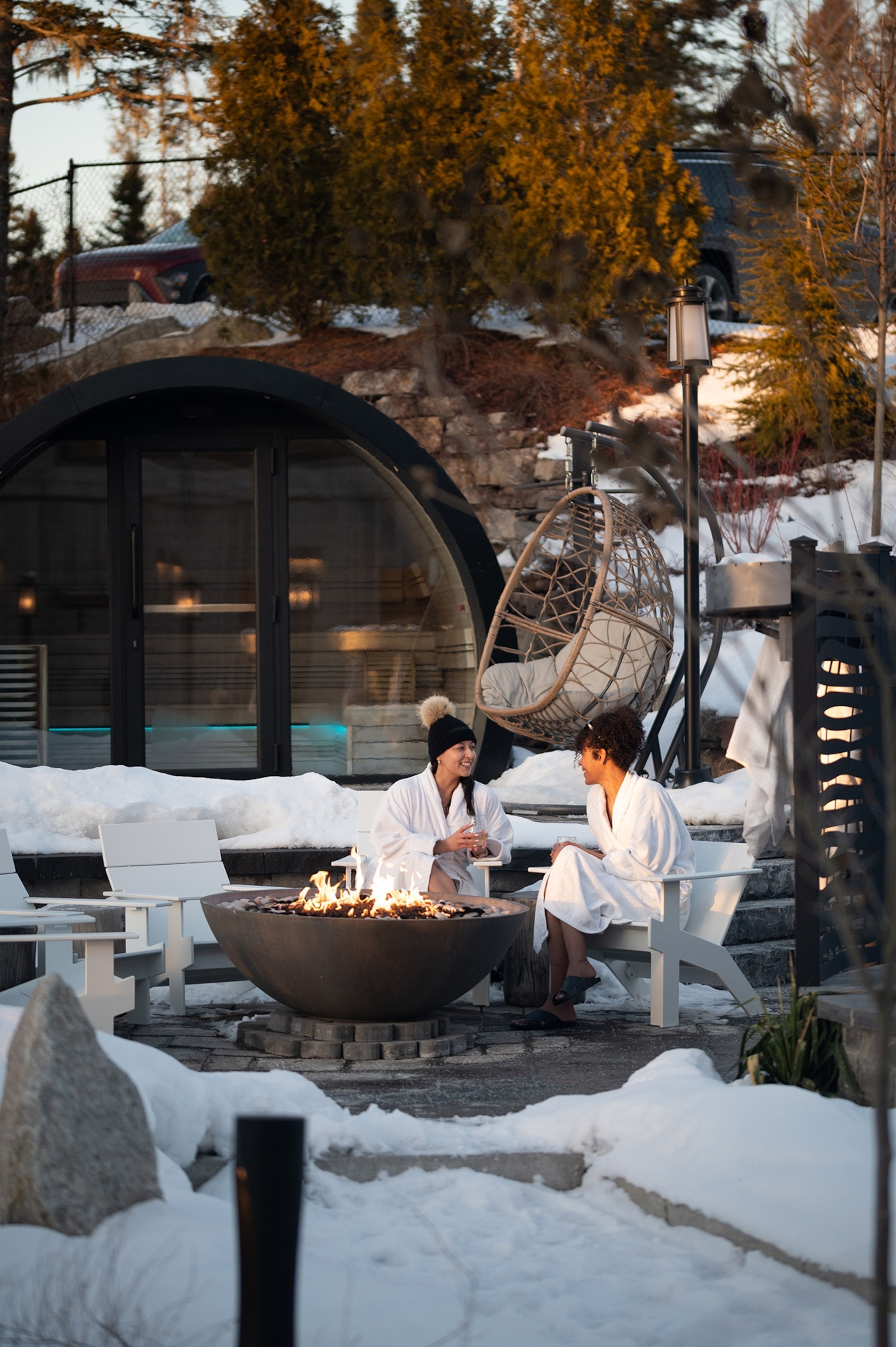 Two women sit chatting on an outdoor sofa in white robes surrounded by a fire pit, sauna building, more seating
