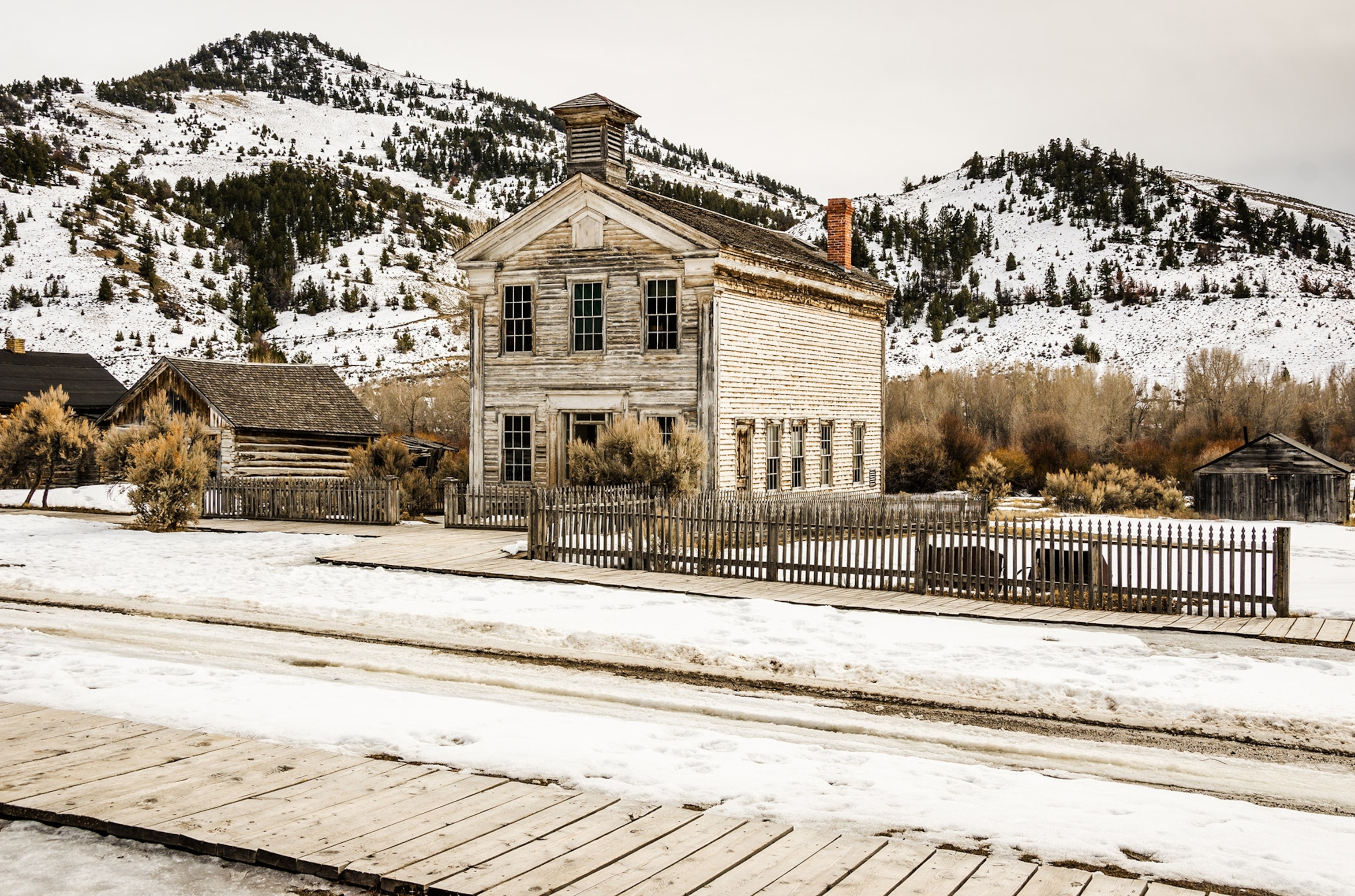 A view across a wooden walkway toward a large white-washed building flanked with two smaller ones, deserted area, no people. Low, snow-covered mountains rise in background.