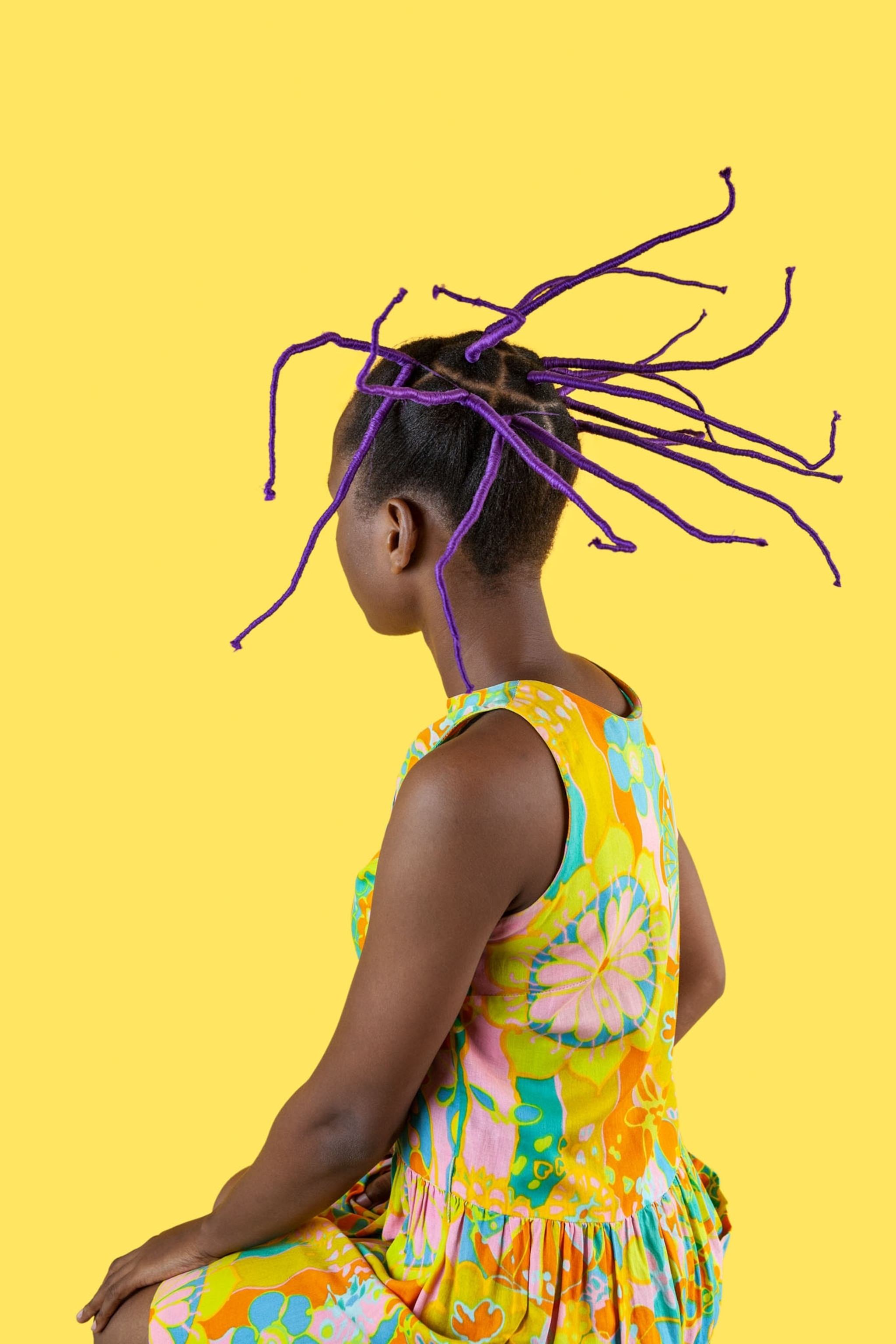Picture of woman in yellow dress with hair knotted with purple thread, on yellow background.