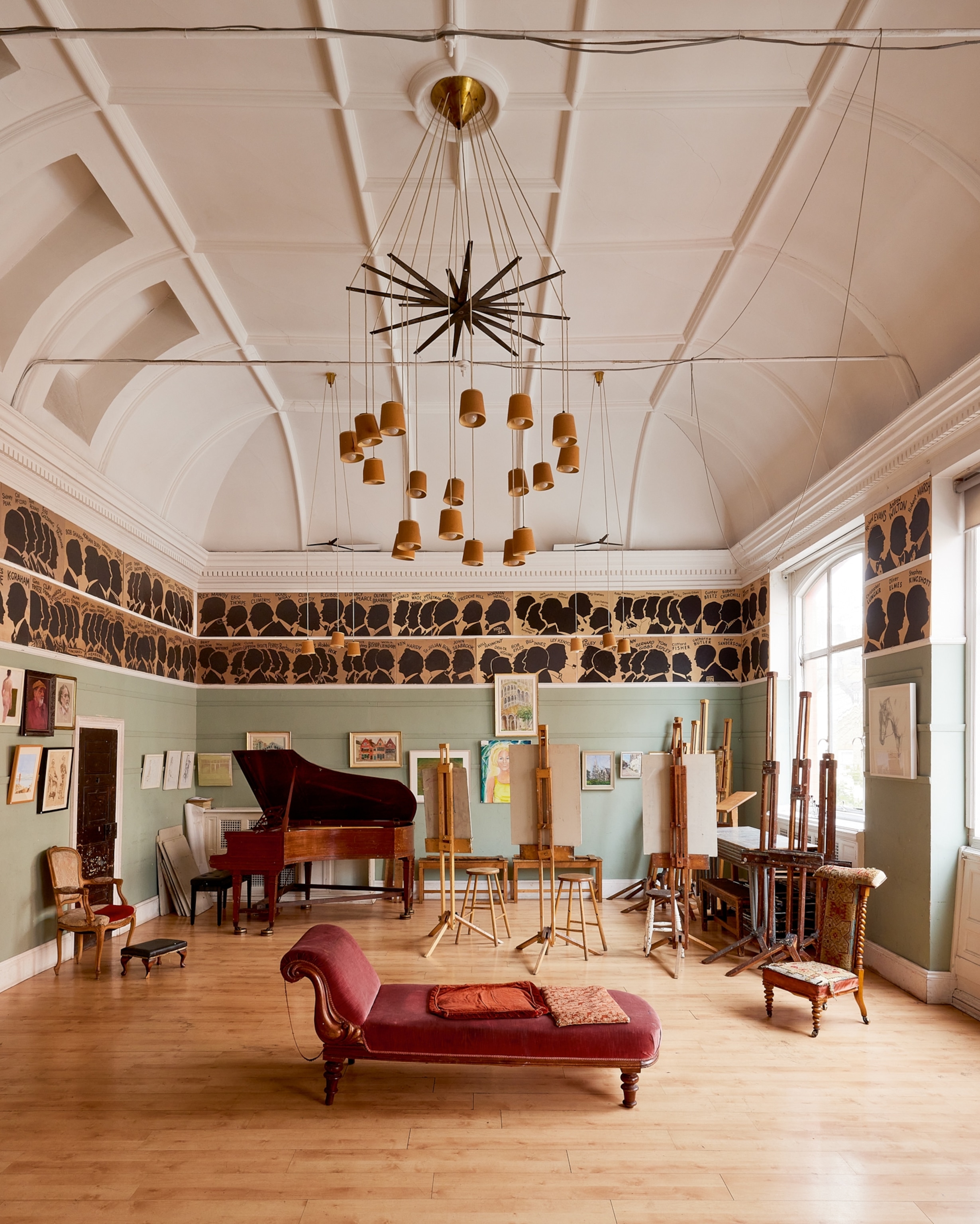 Spacious art studio with vaulted ceiling, featuring a grand piano, easels, and a red chaise lounge. The mood is creative and elegant.