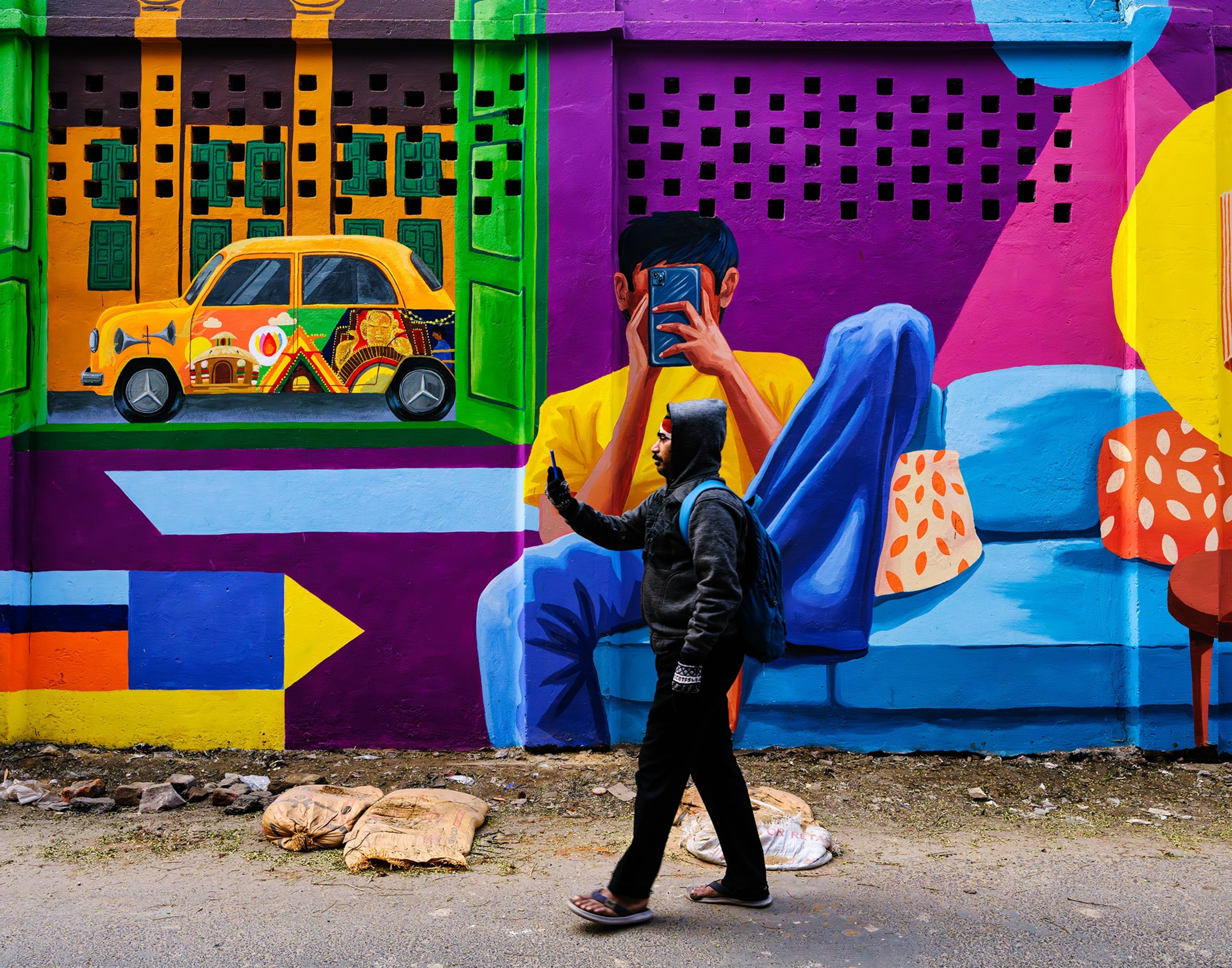 A young man pausing on a street in Kolkata to take a picture as a mural behind him shows a man taking a photo, too.