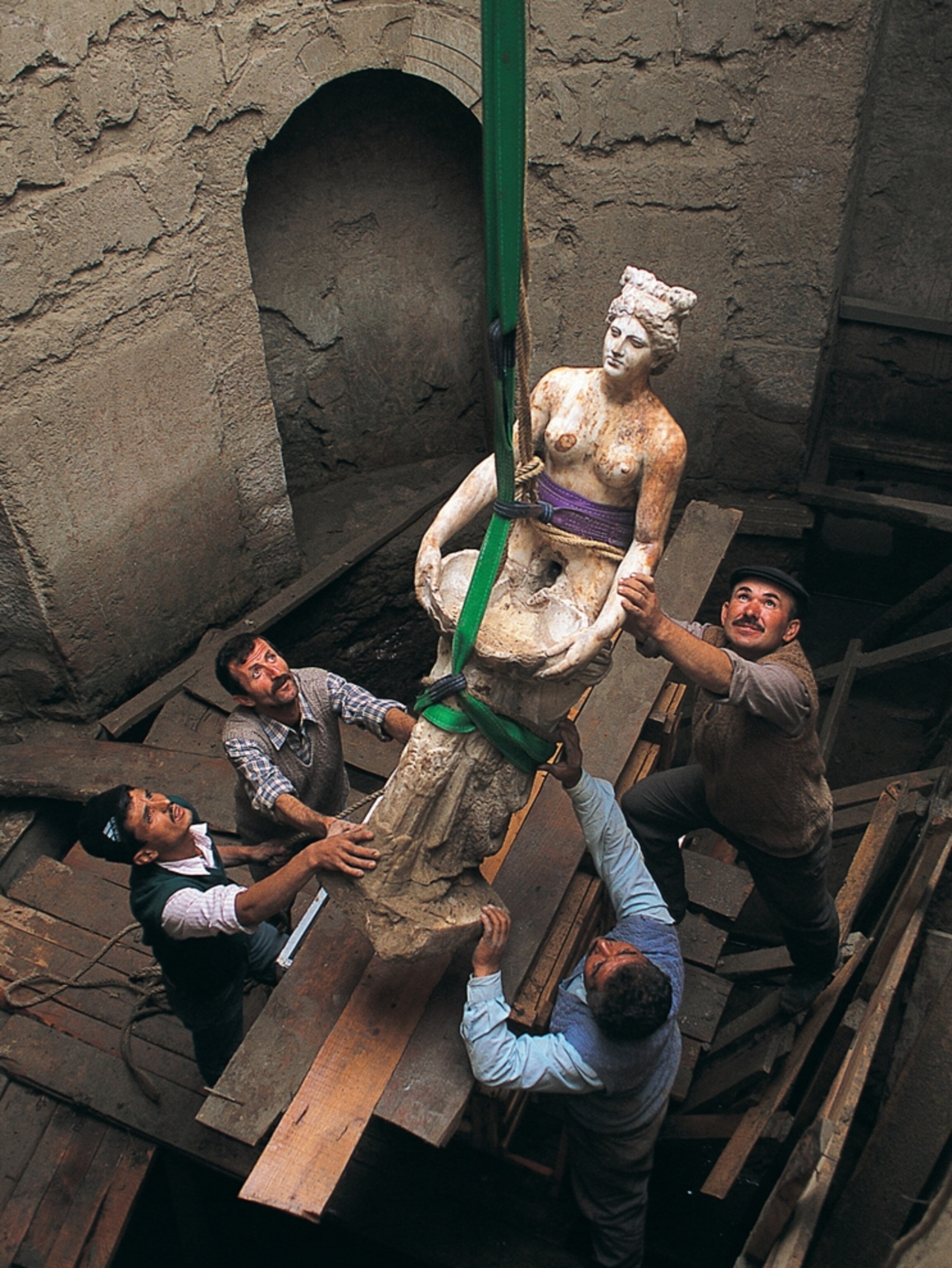 A Roman statue of a bathhouse nymph is lifted to the surface by four men and some colorful straps.