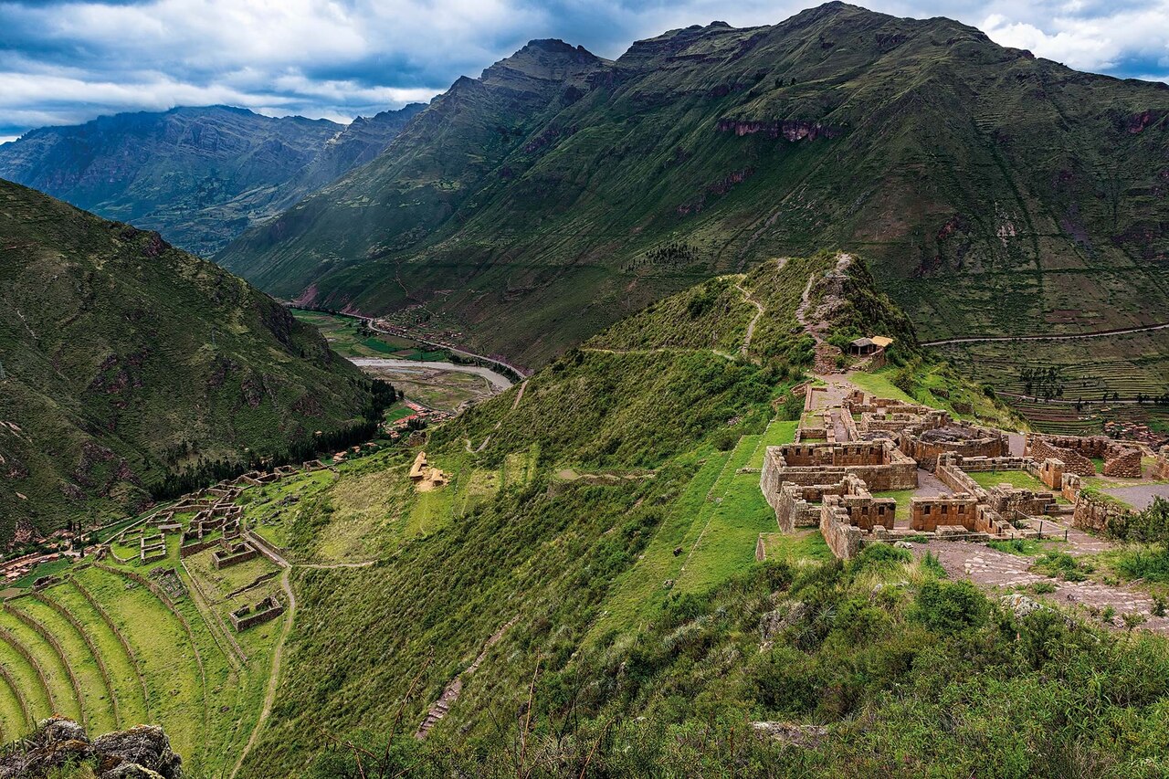 A complex of ancient stone buildings overlooks the Sacred Valley of the Inca from a hilltop, with mountains in the background