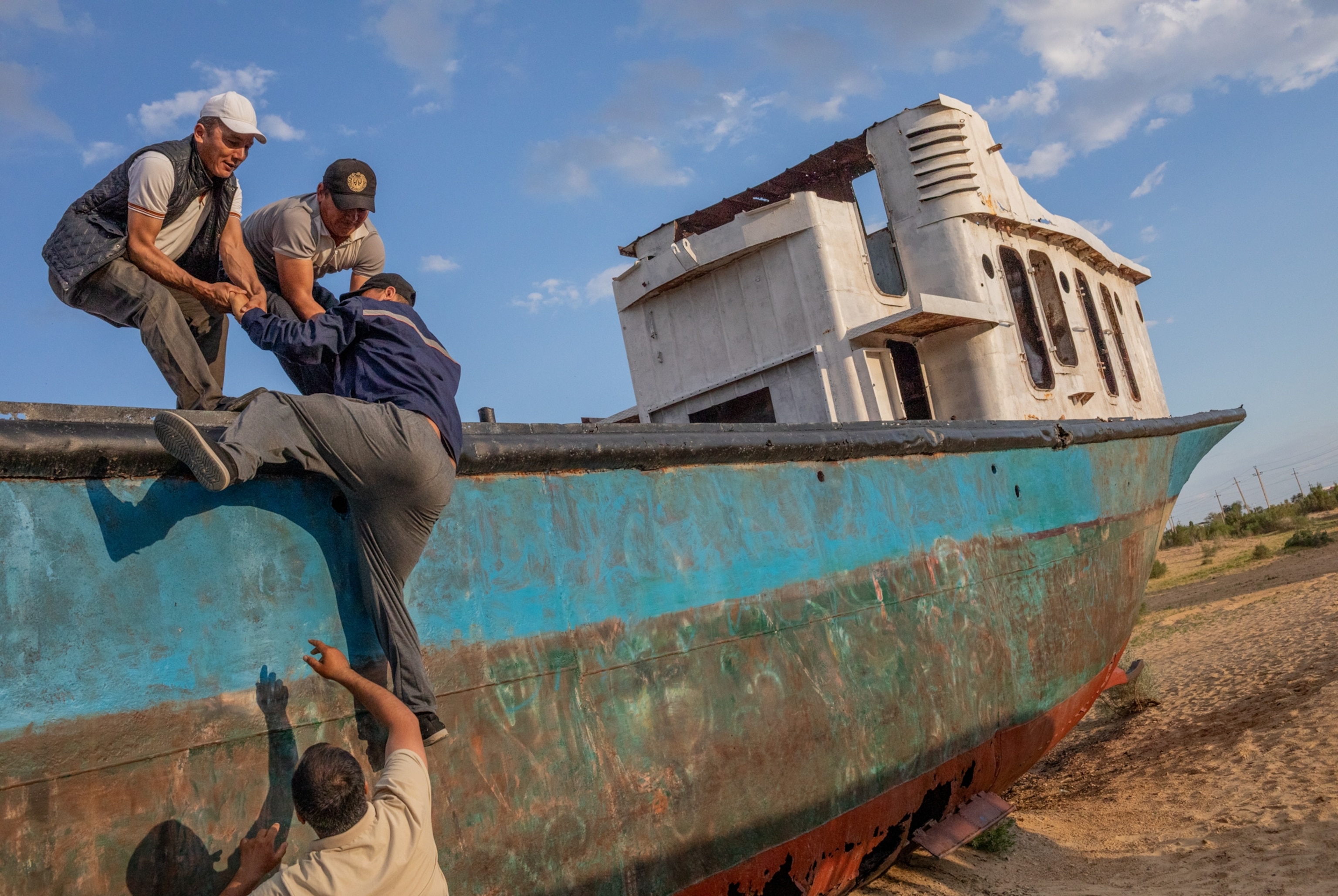 Four men helping each other to climb on rusty once torque colored boat sitting on dry seabed.
