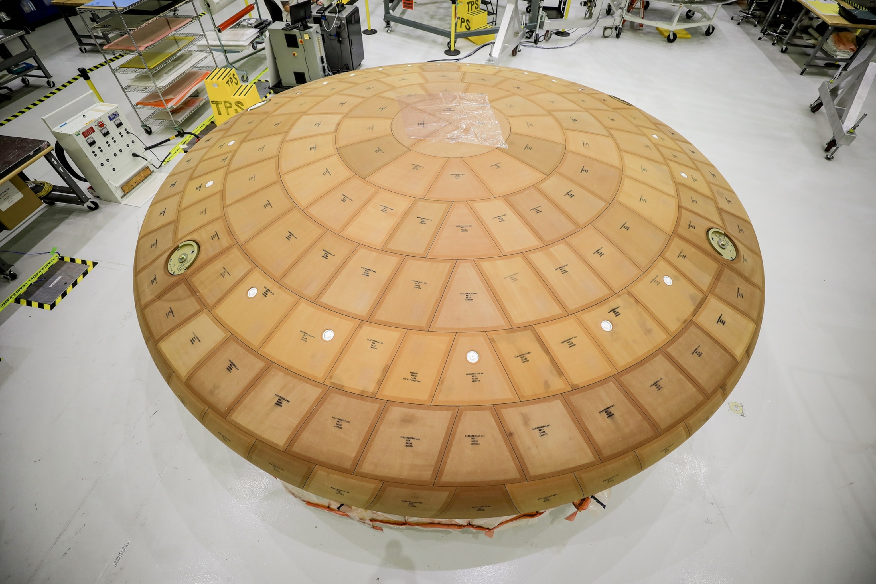 A large, circular heat shield with a grid of tan panels lies on a clean laboratory floor