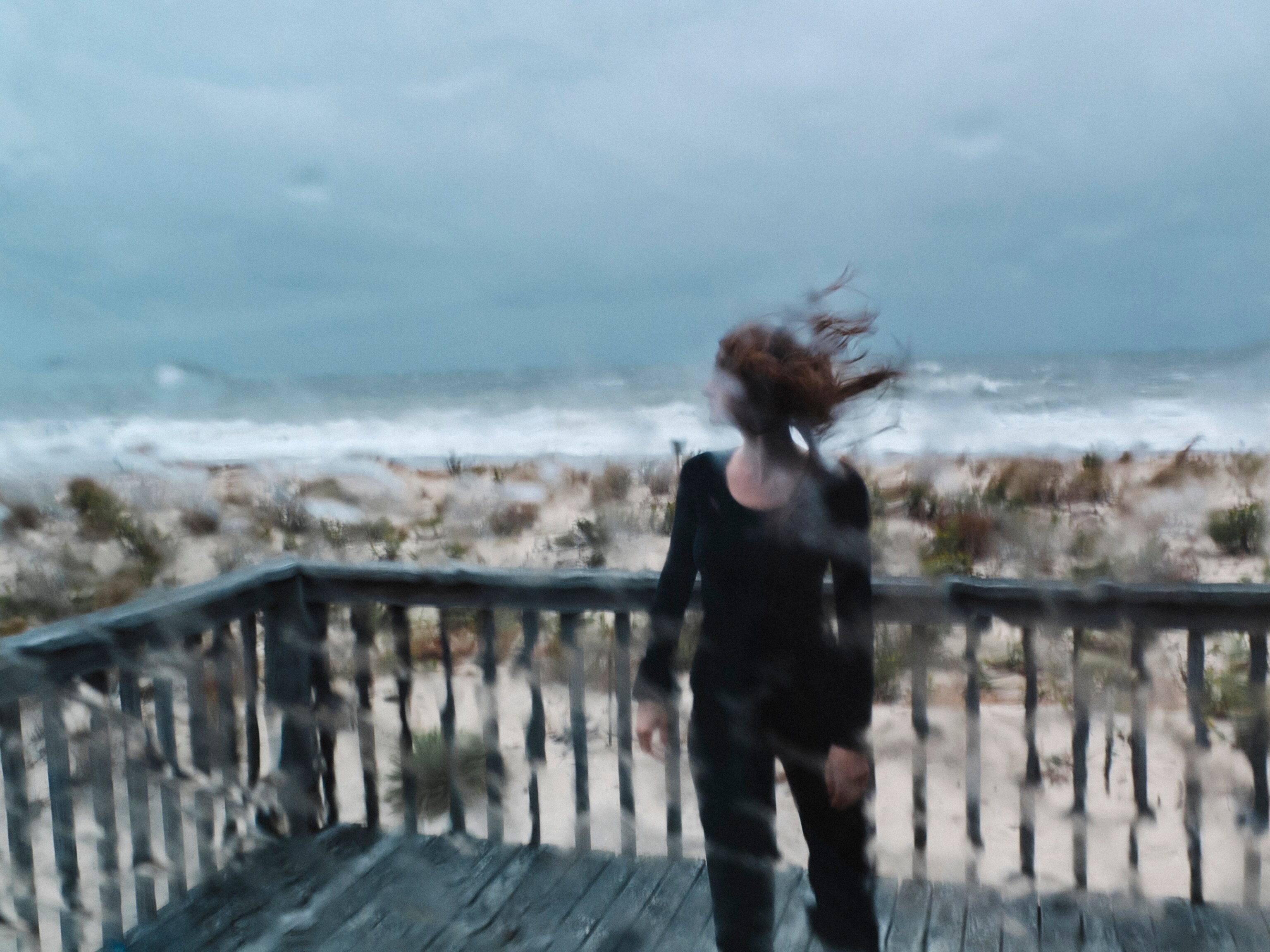 a woman with long brown hair blowing in the wind standing on a deck during a storm, shot through glass with rain on it