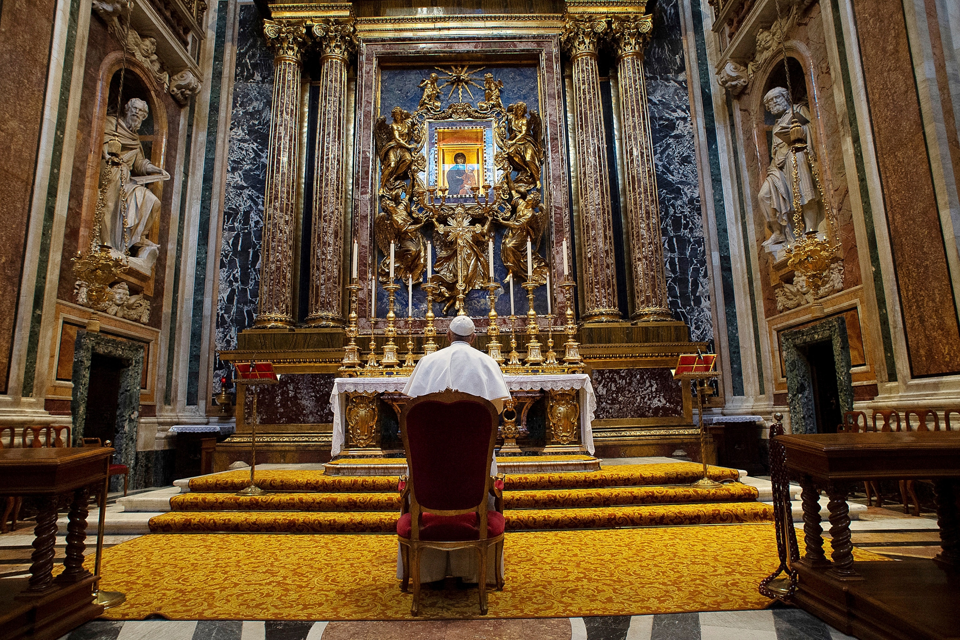 Pope Francis stands with his back to the camera as he prays in front of a large altar.