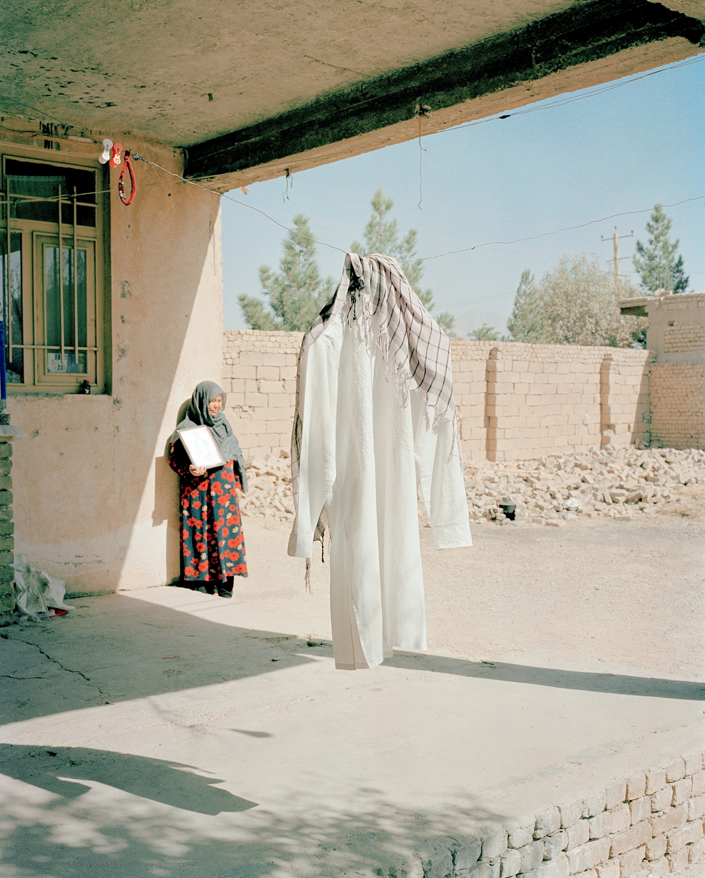 A mother stands by the wall of her home while the robe of her late son hangs on a line in front of her.