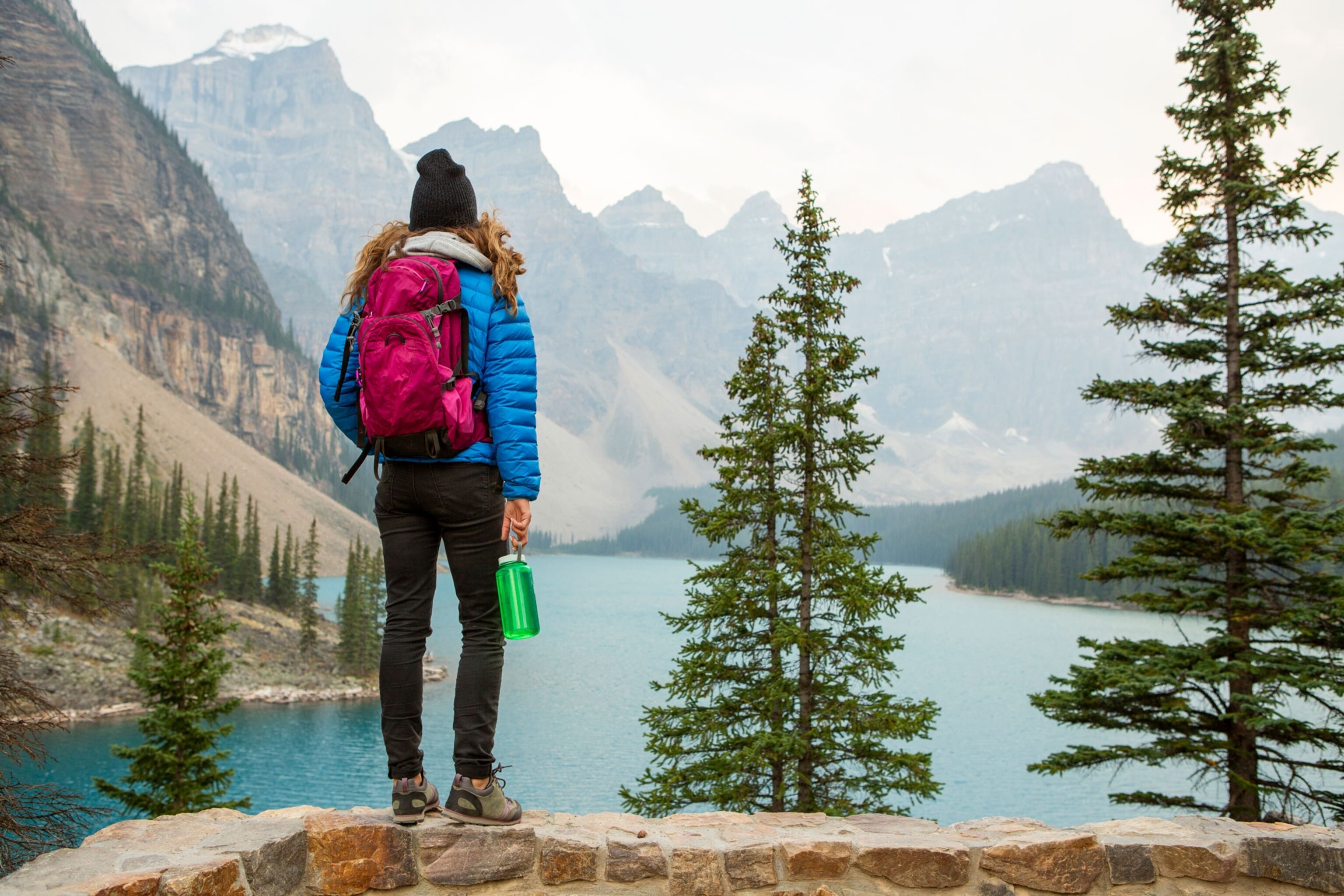 A woman with a red backpack, blue coat, black pants and hiking boots hiking around a blue Lake in the mountains