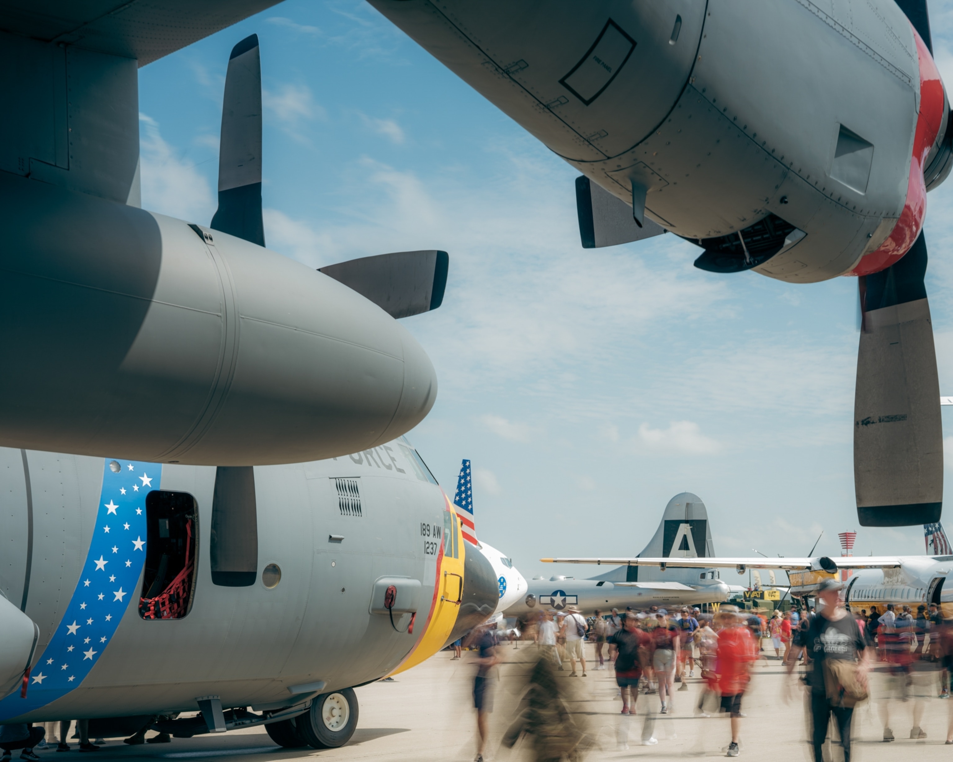 Aircraft display at an air show, featuring large propellers and fuselages of military planes. Crowds of blurred people explore under a clear sky