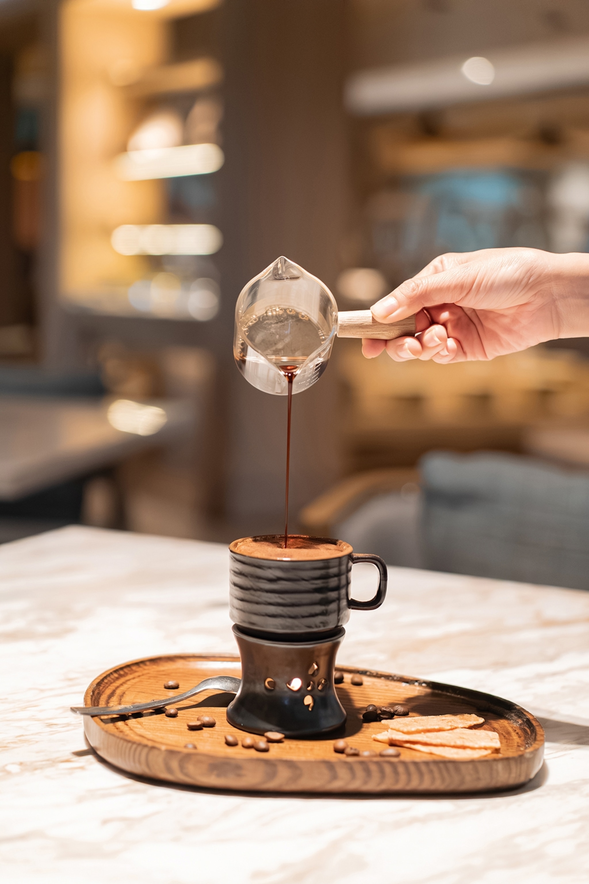 A hand pouring coffee into a small, elevated espresso cup from a glass steamer cup.