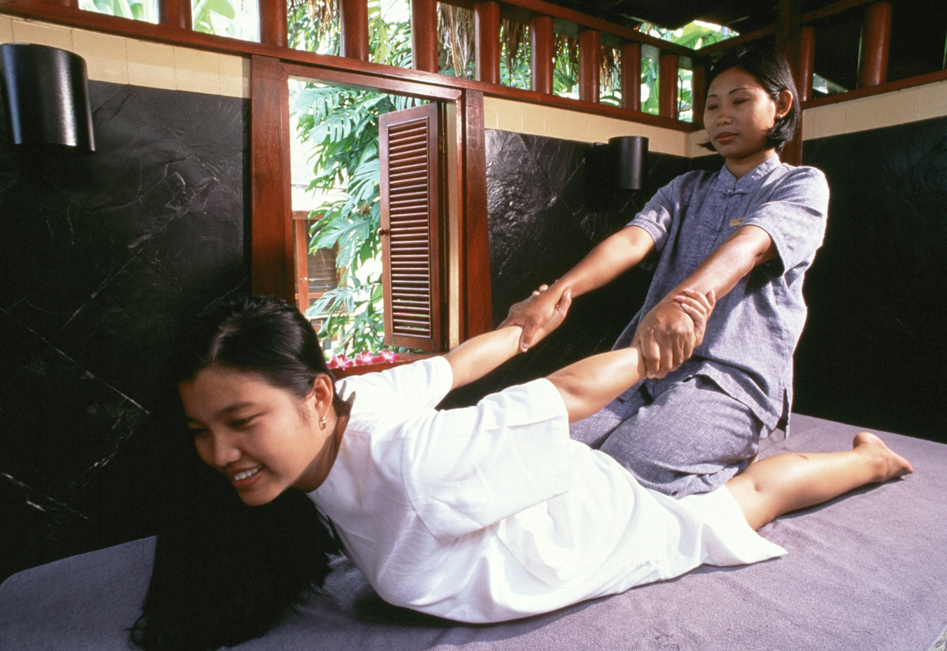 A woman in a large white t-shirt dress is laying on her stomach with her arms back while another woman is kneeling behind her and pulling her arms.