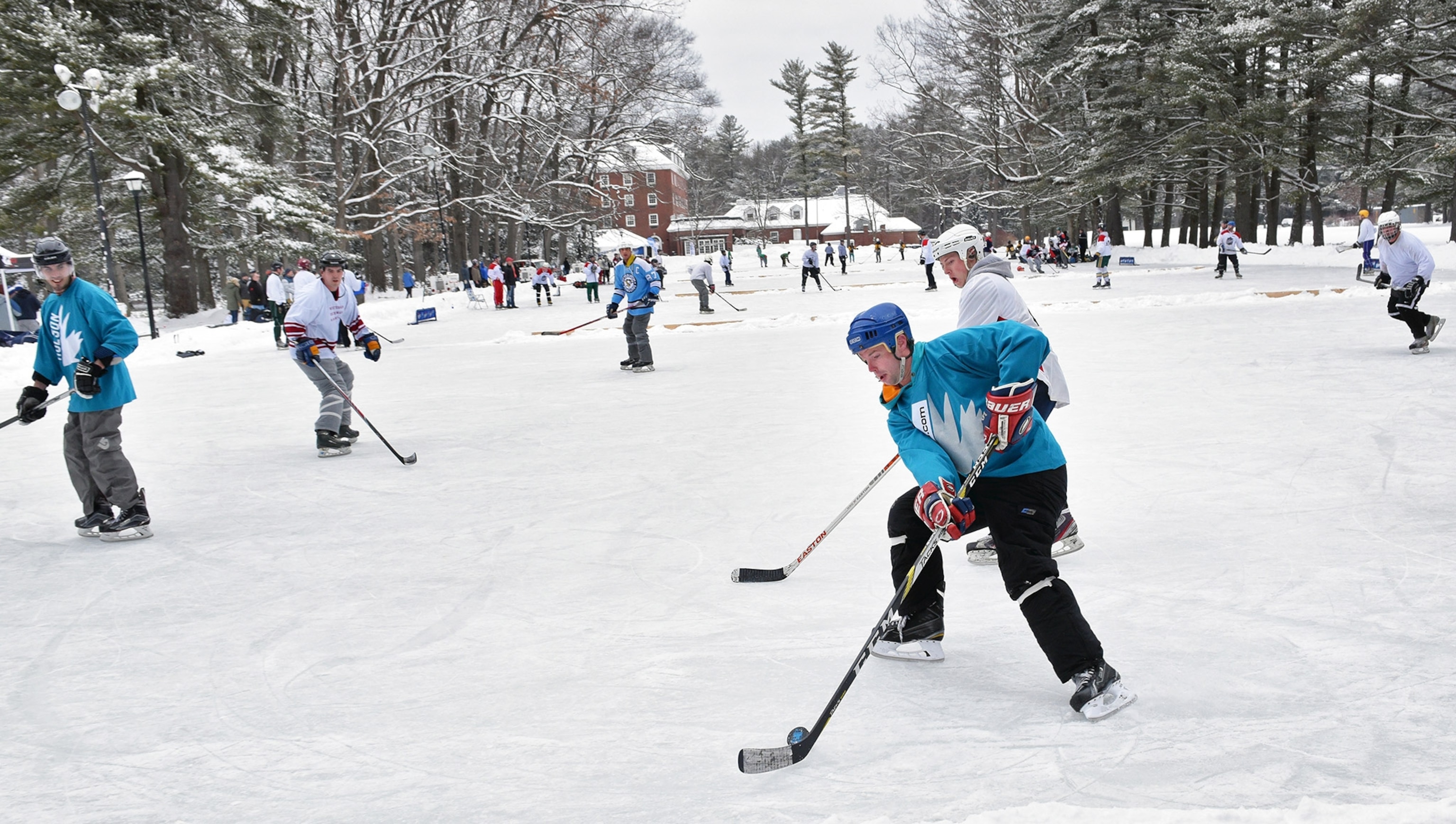A group of young men in hockey uniforms and skates play a game on a stretch of ice, trees and historical buildings in the far background