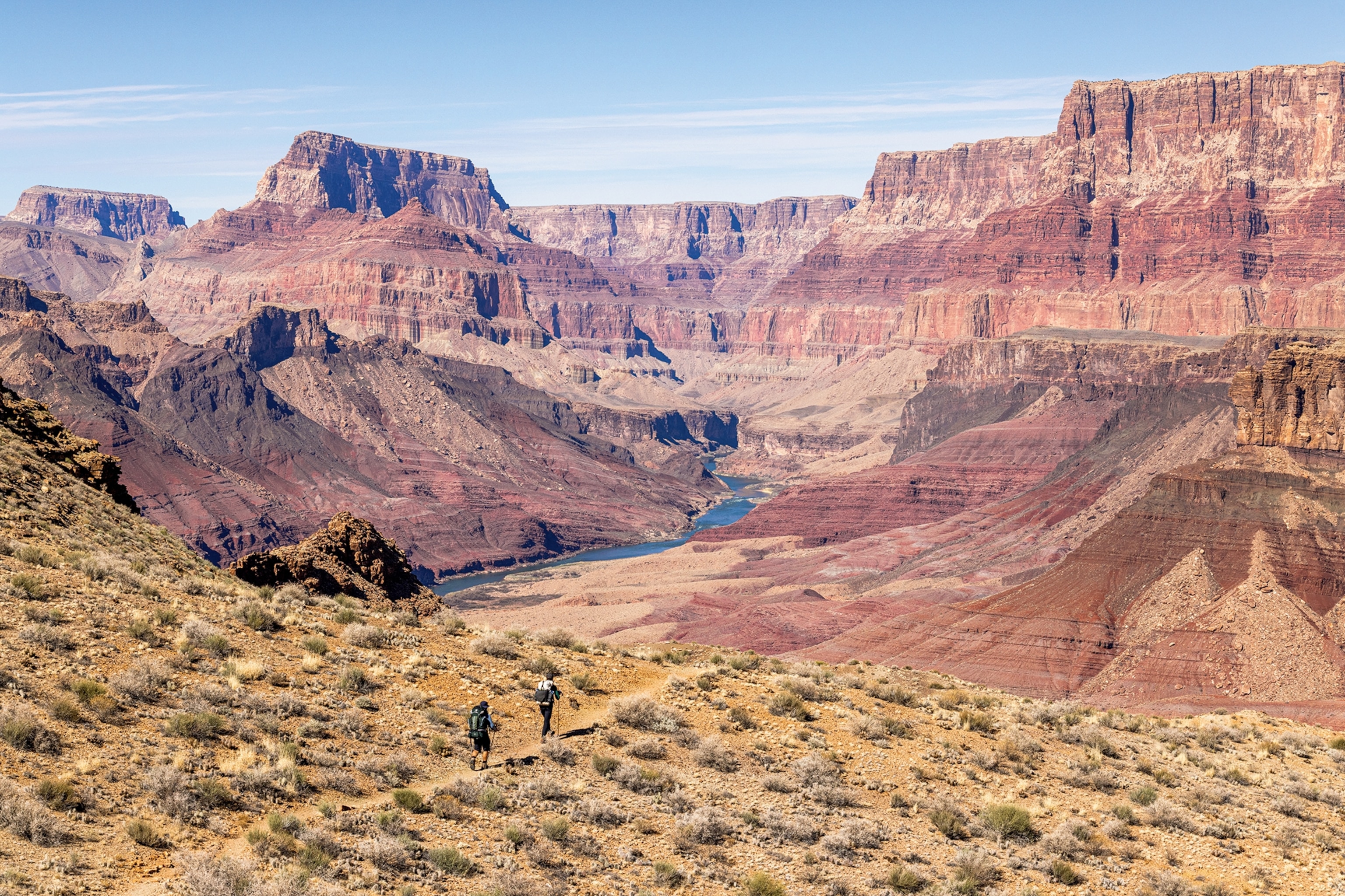 An aerial shot of two hikers walking down a stony trail with the impressive landscape of a canyon in front, and a river snaking through the bottom.