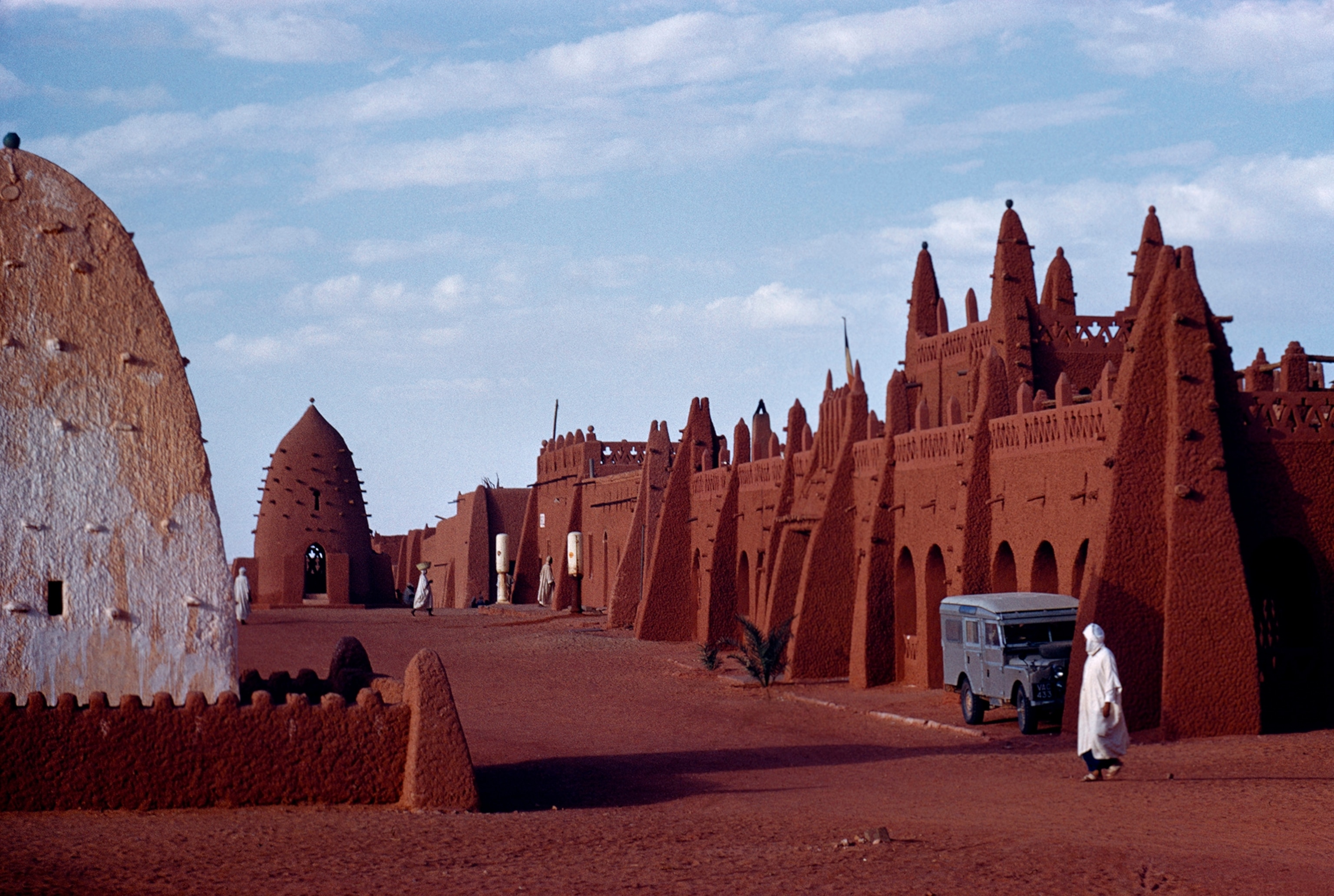 This view shows reinforcing beams protruding from adobe walls, tomb rises at the end of the street. The French Earthen fence on the left surrounds the whitewashed Tricolor surmounts a hotel. Land-Rover and gasoline pumps offer modern contrast.
