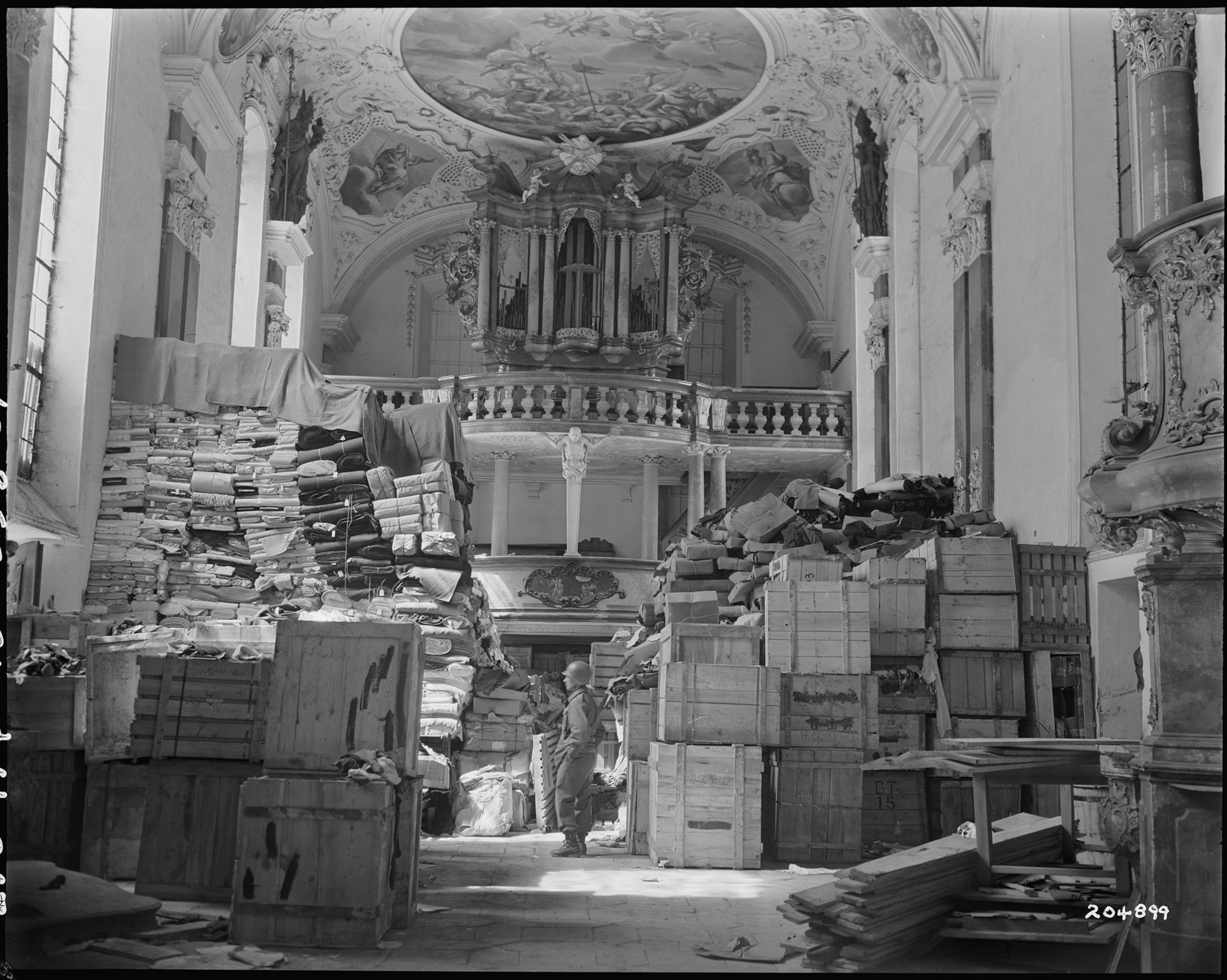 A black and white photogrpahshows a man in uniform with stacks of boxes, pallets, and paper surrounding him.