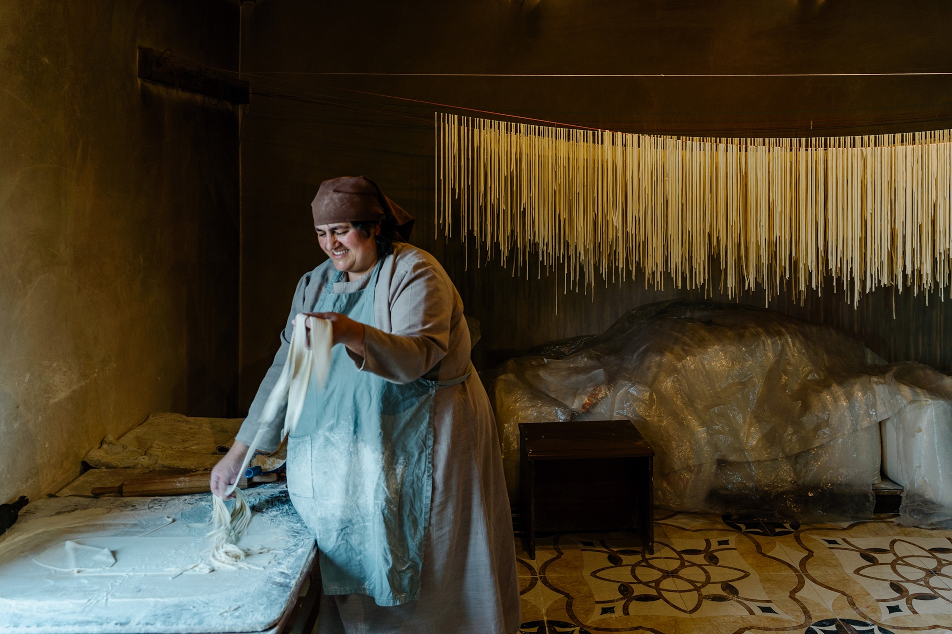 A middle-aged woman wearing a simple chef apron in a dark, rustic kitchen, pulling pasta by hand and hanging them up on a rod behind her to dry.