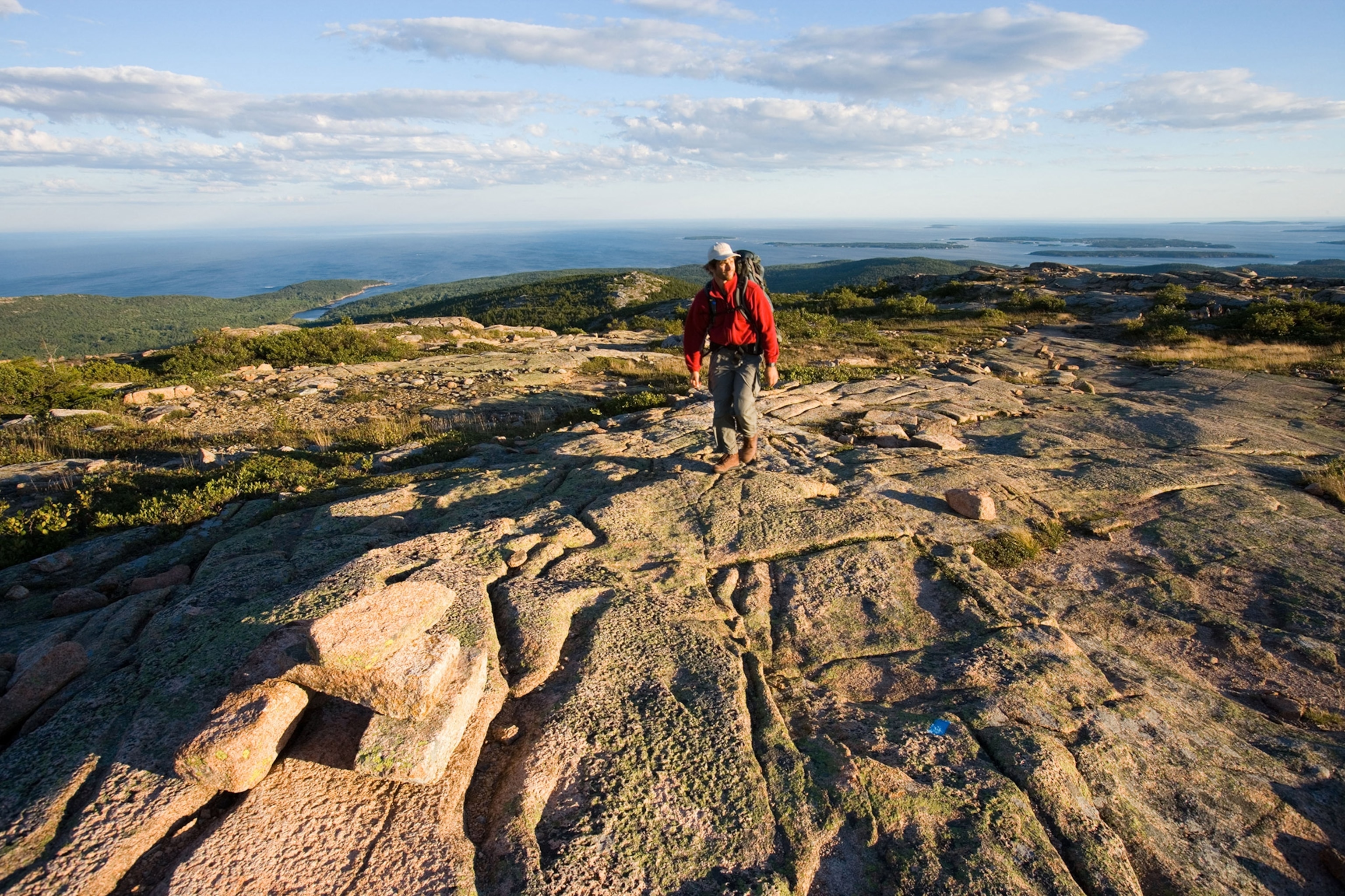 A man in a red jacket walks across a high, flat rocky plateau, ocean in the distance