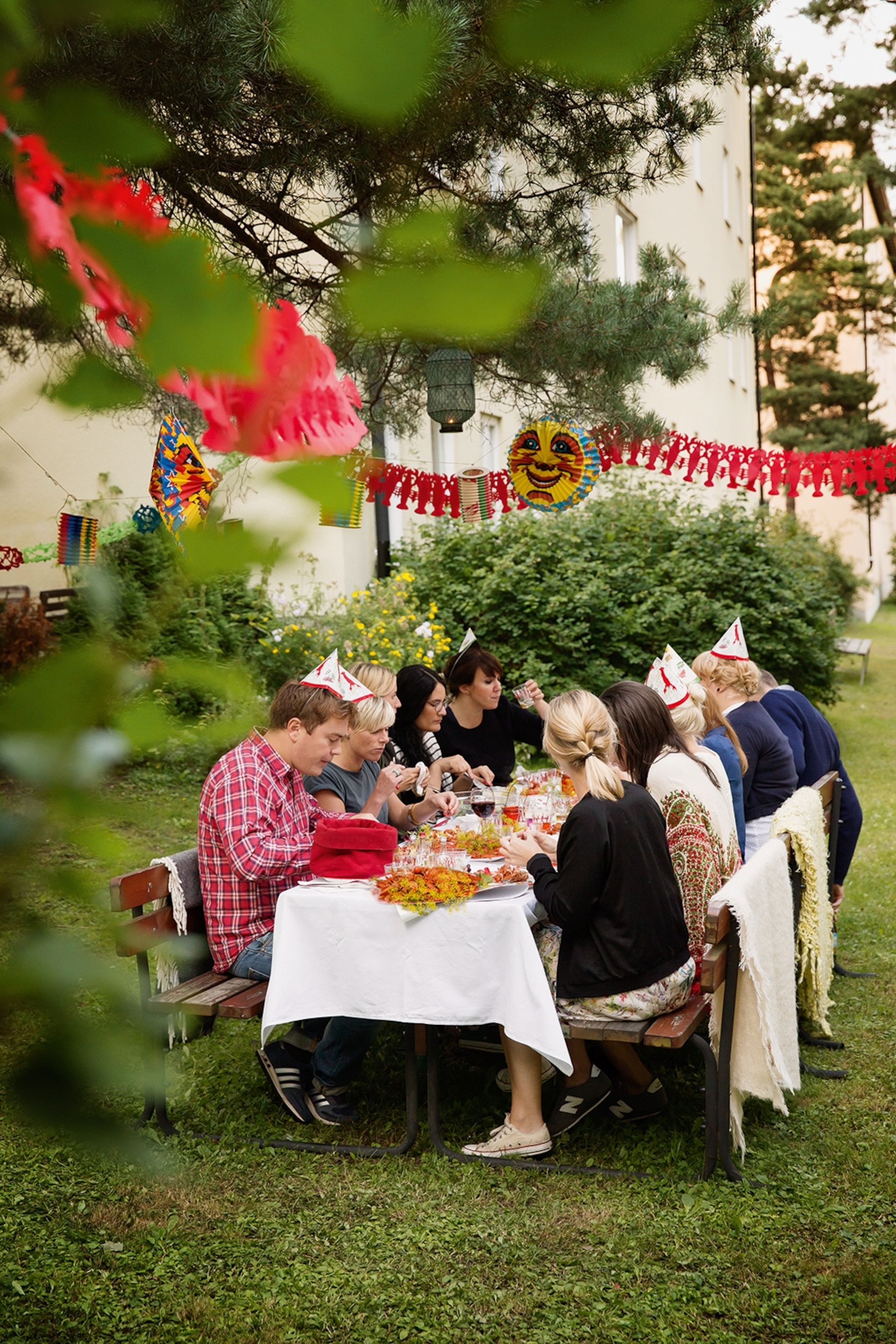 A group of diners wearing party hats, sat at a table, eating crayfish in a garden decorated with bunting.