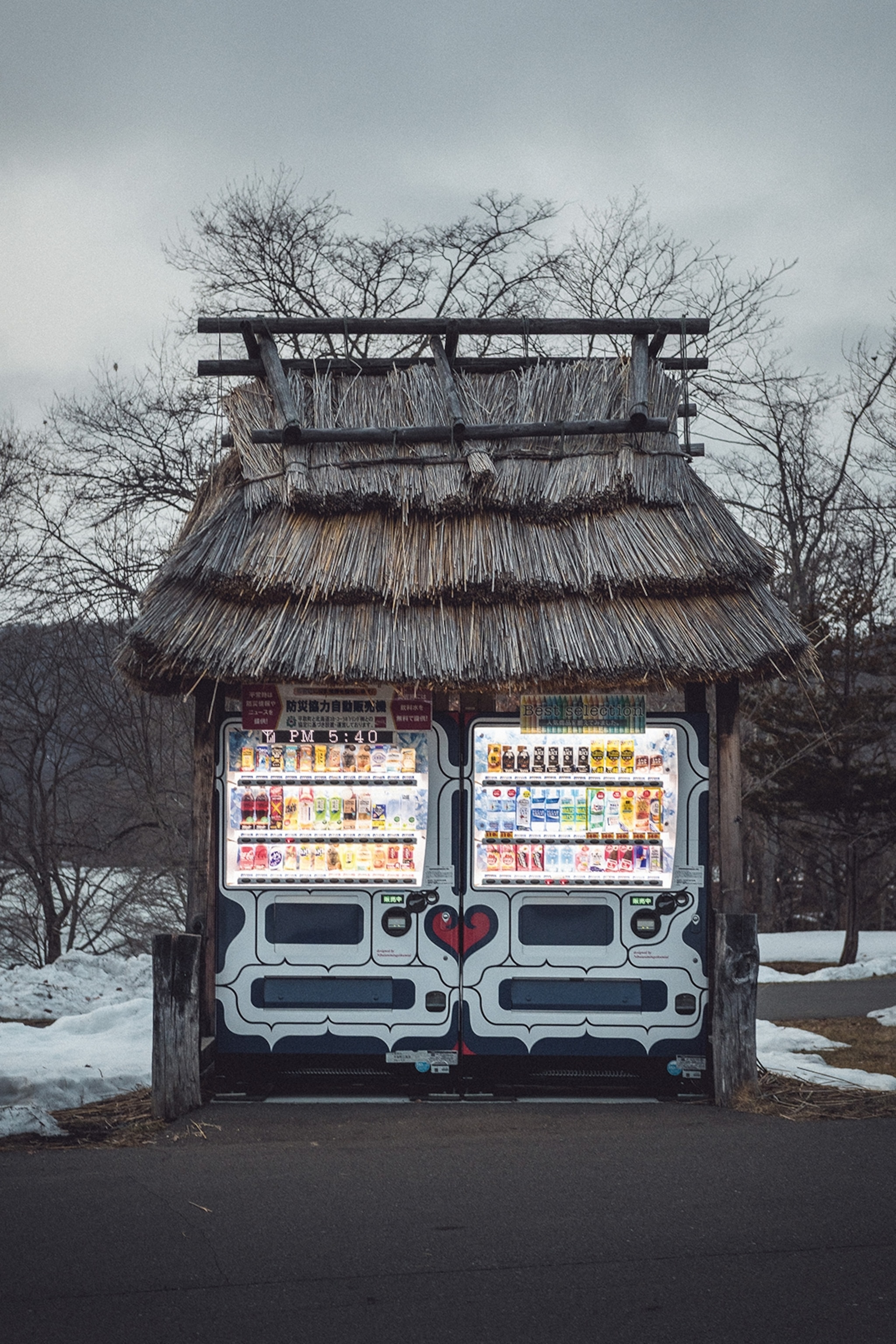 A snowed-in vending machine with a straw roof on the side of a road at late dusk.