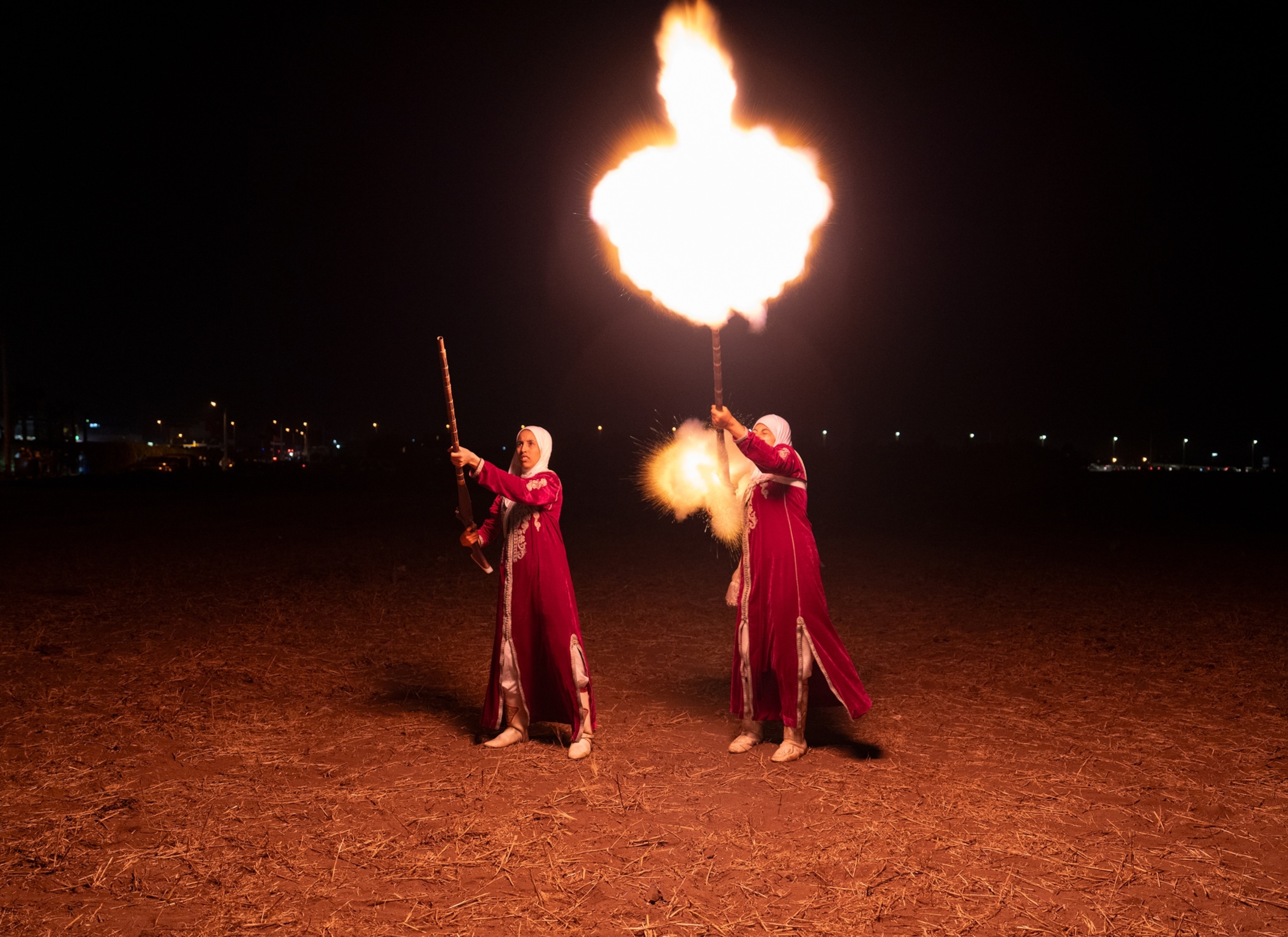 Two women wearing red stand in the dark and fire off their guns. The gun on the right emits a large flame in the air that lights up the area around them.