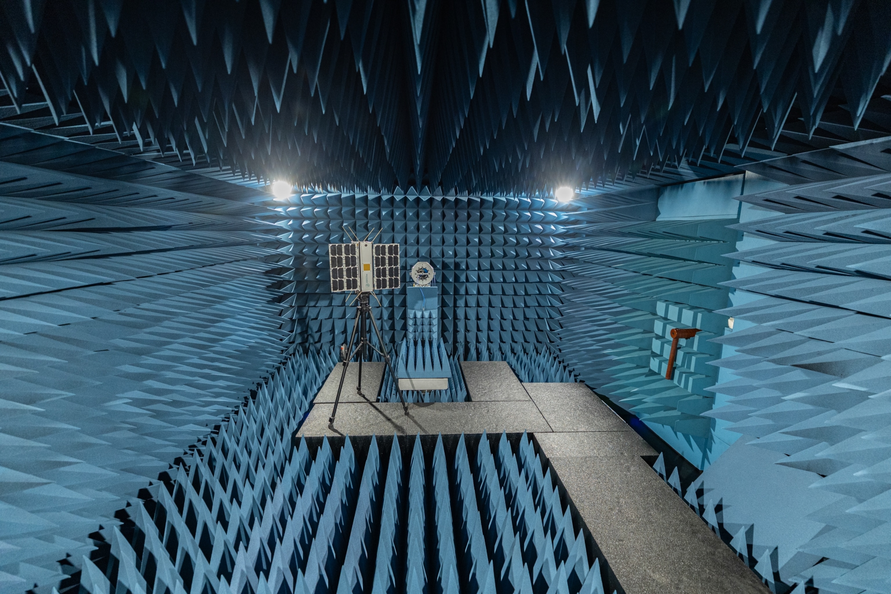 A room with blue, spiky, sound-absorbing walls features a satellite equipment setup on a tripod under bright lights