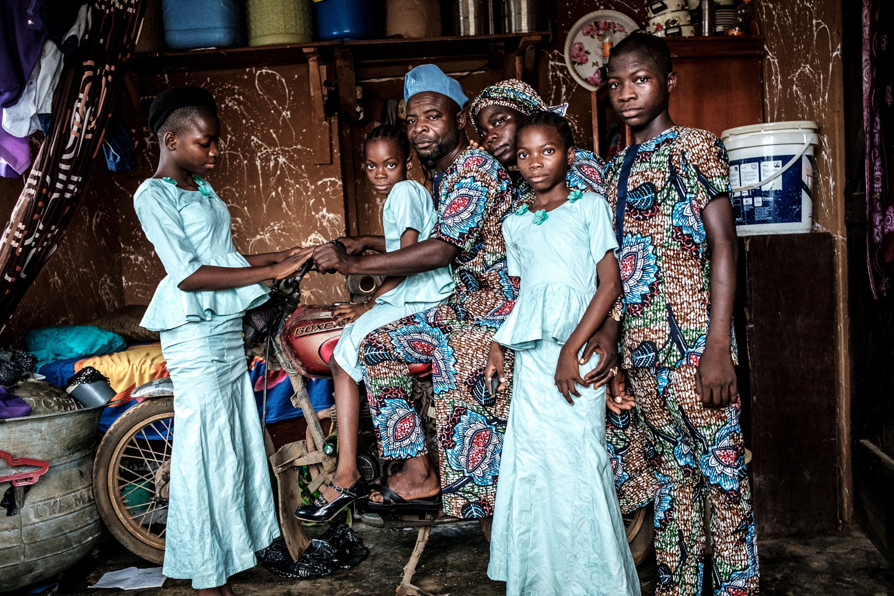Man on his bike inside his one-room apartment with his wife and one daughter dressed in the same fabric as he, and three other girls dressed in pale-blue long dresses.