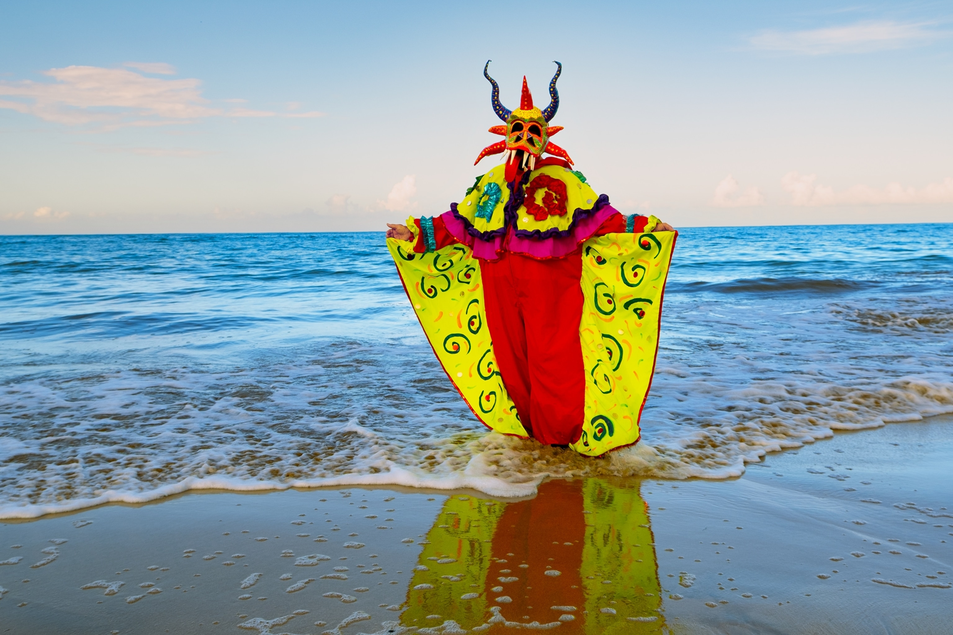 A person wearing a colorful horned mask and bright ceremonial costume stands in shallow ocean water at the edge of a sandy beach, with gentle waves and a clear blue horizon in the background.