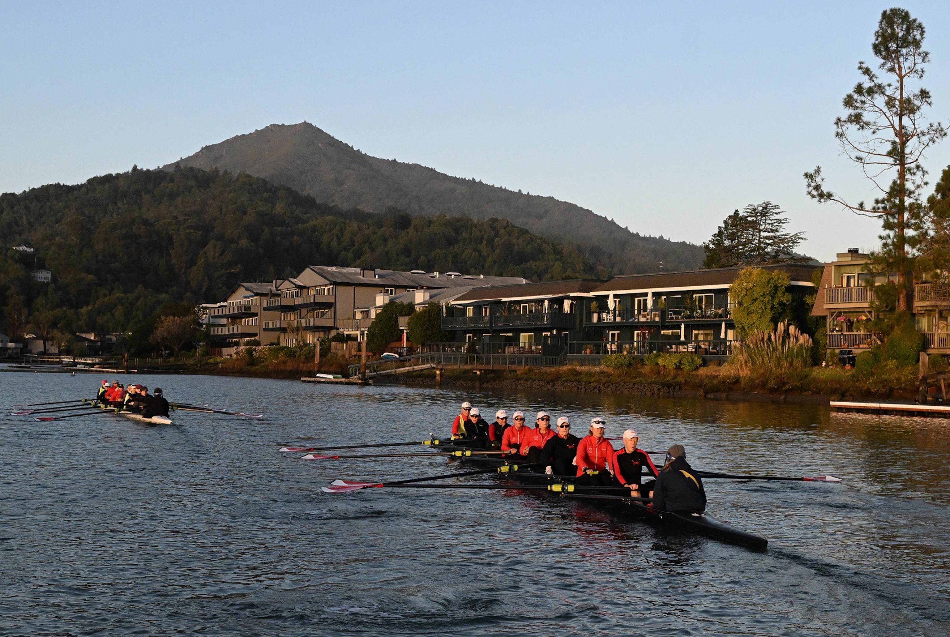 A row boat of women in red jackets in a lake with mountains and houses in the background.