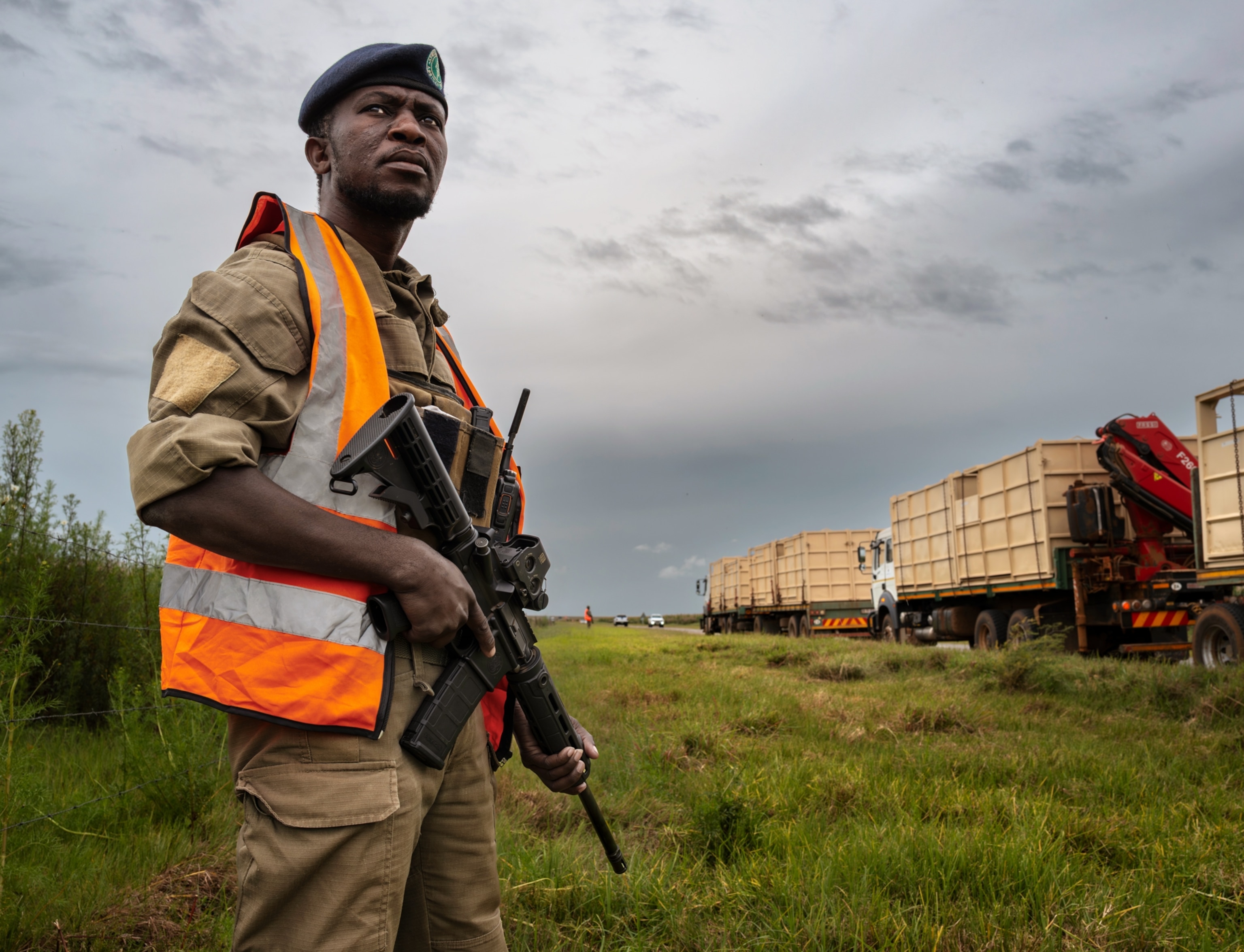 A man wearing a tan uniform and an orange vest stands in a. grass field with a large gun as trucks with large metal containers drive on the road next to him.