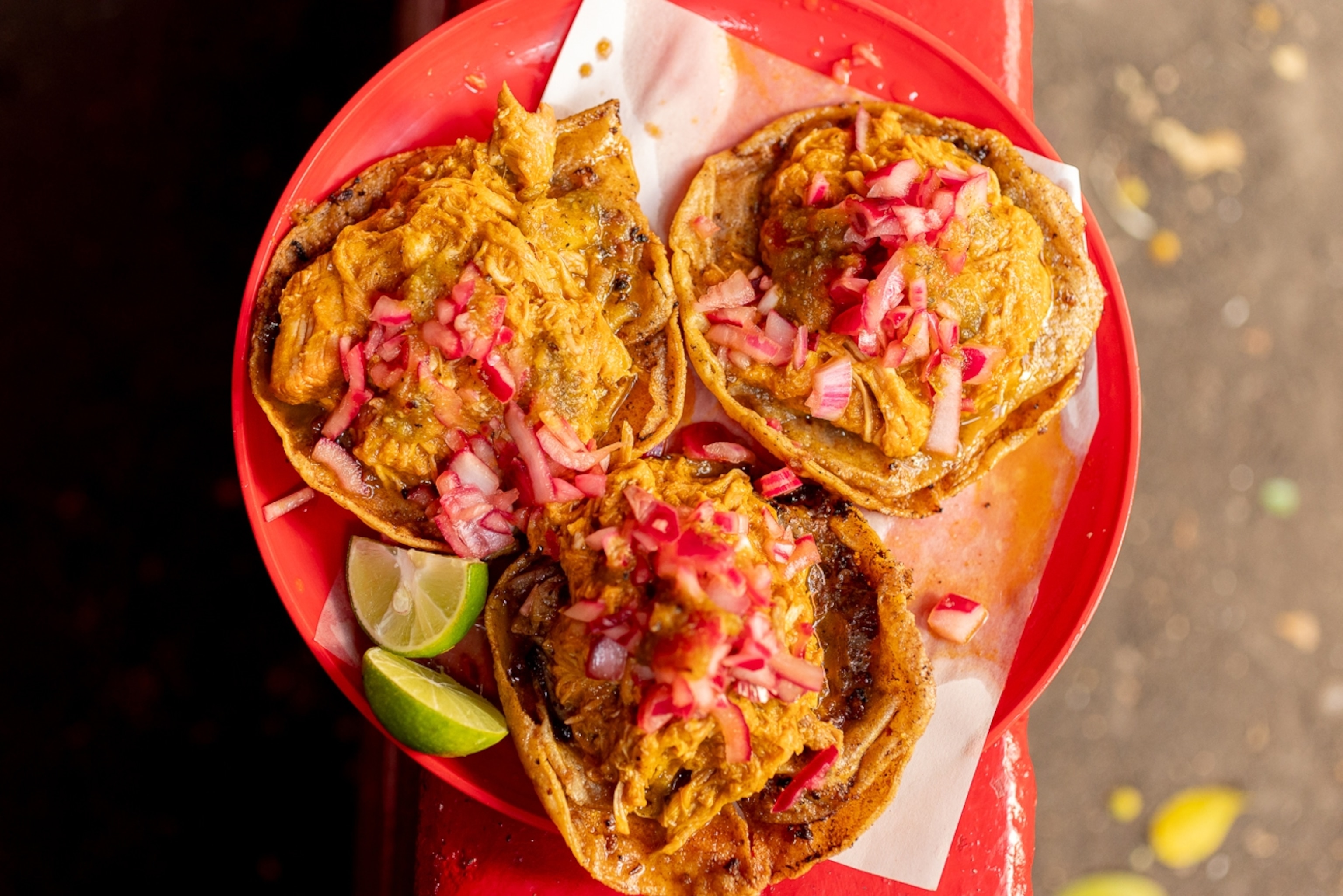A close-up of a plate of three pulled pork tacos topped with pickled red onion and lime wedges.