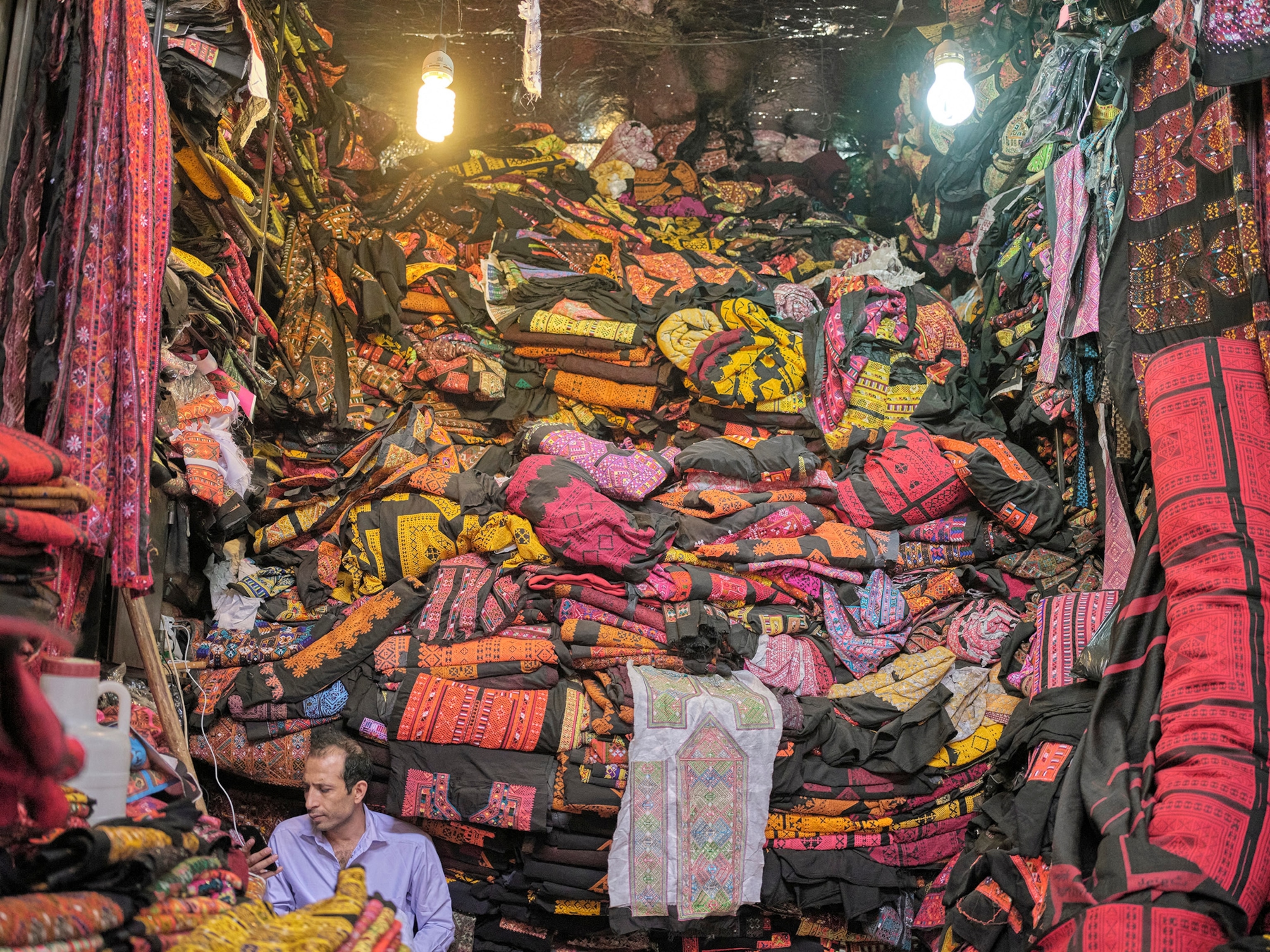 A man in a purple shirt sits in front of towering pile of pink, red, orange, yellow, black fabrics, folded.
