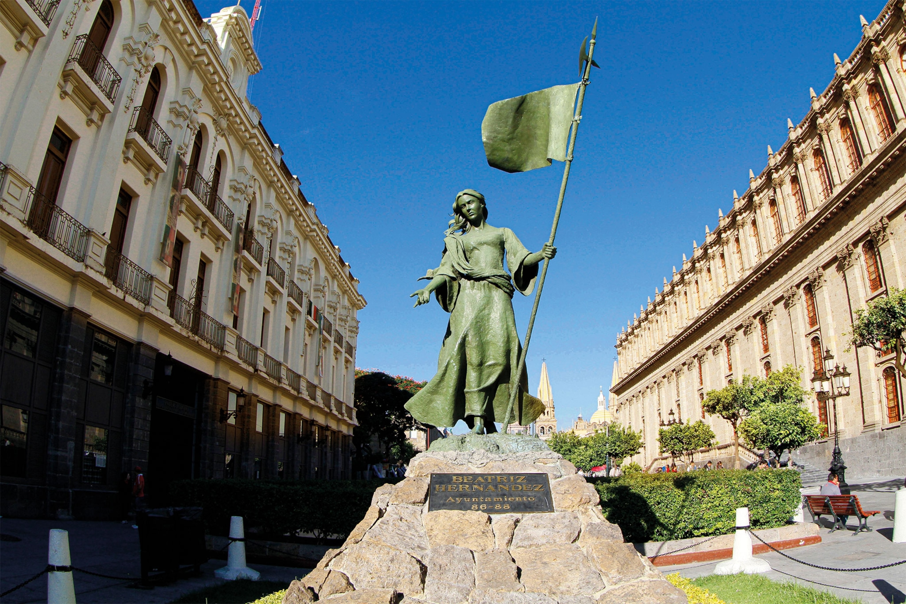 A bronze statue of Beatriz Hernández holding a flag stands near a city plaza
