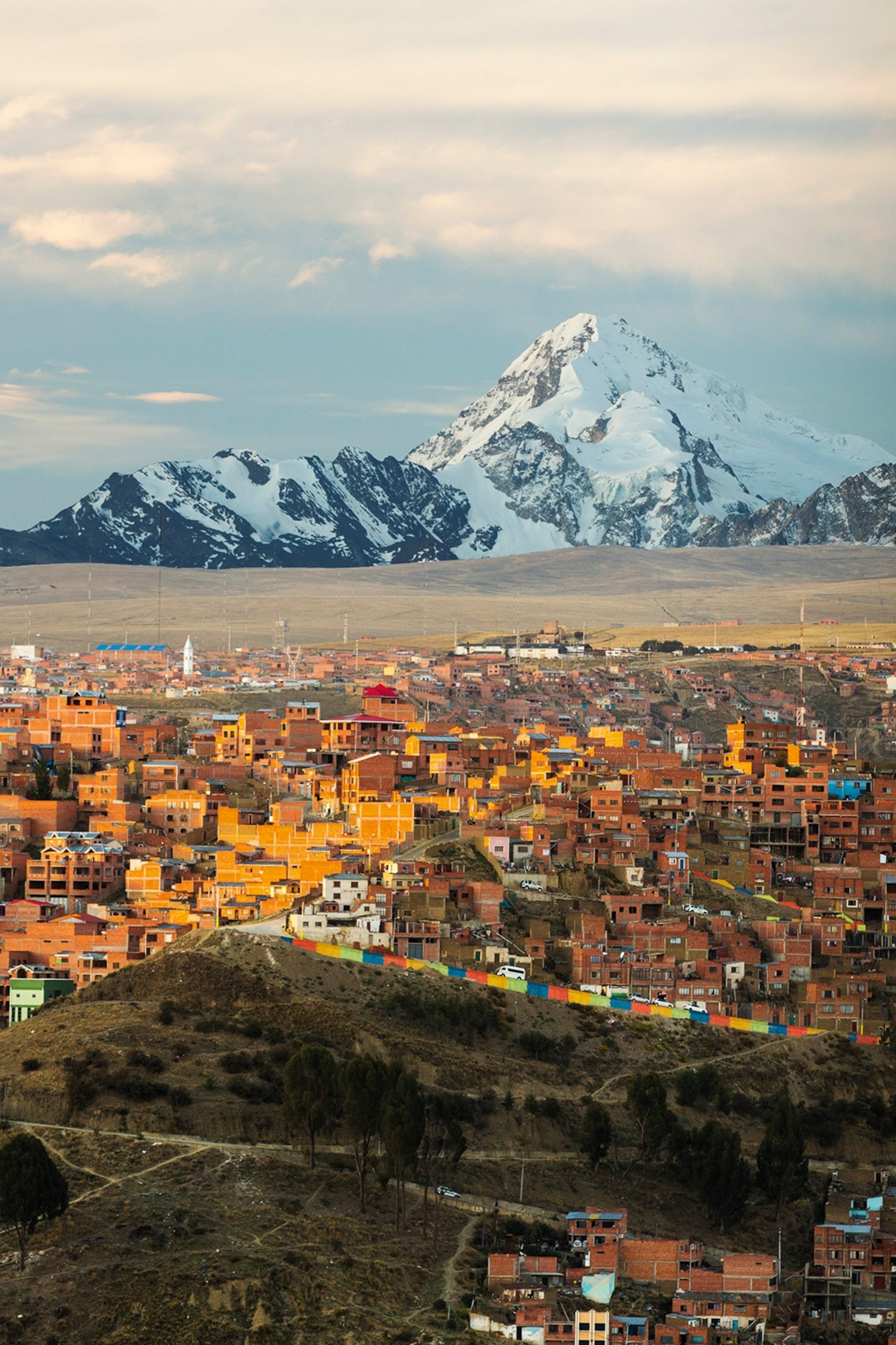 A town seen from the distance with stone buildings catching the sunlight and snow-capped, Andean peaks in the background.
