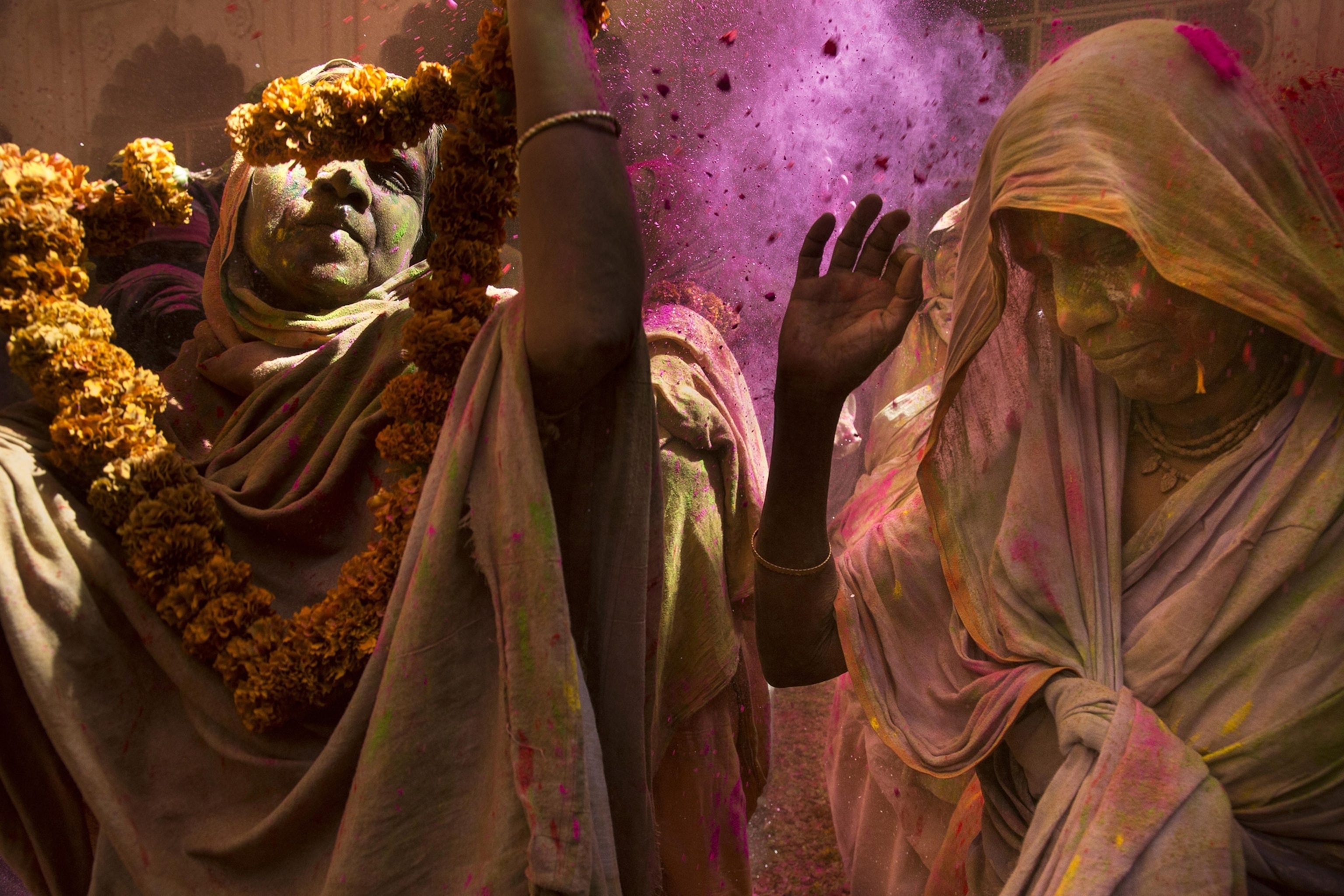 Two women in head scarves and with floral garlands and covered in colorful powder raise their hands in celebration
